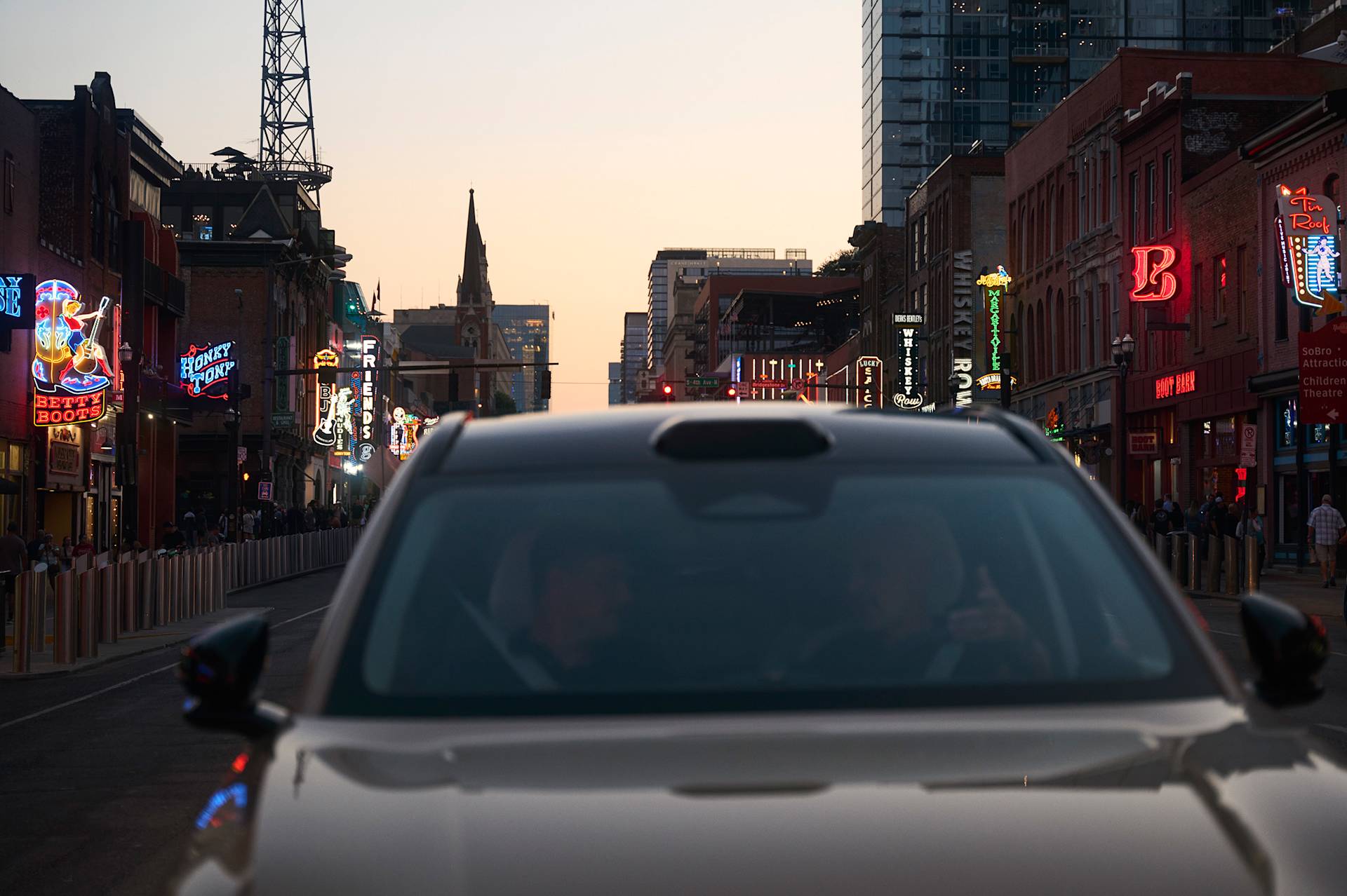 A car in the foreground with two people inside, driving down a lively city street lined with colorful neon signs and tall buildings at sunset. Crowds walk along both sides of the street.