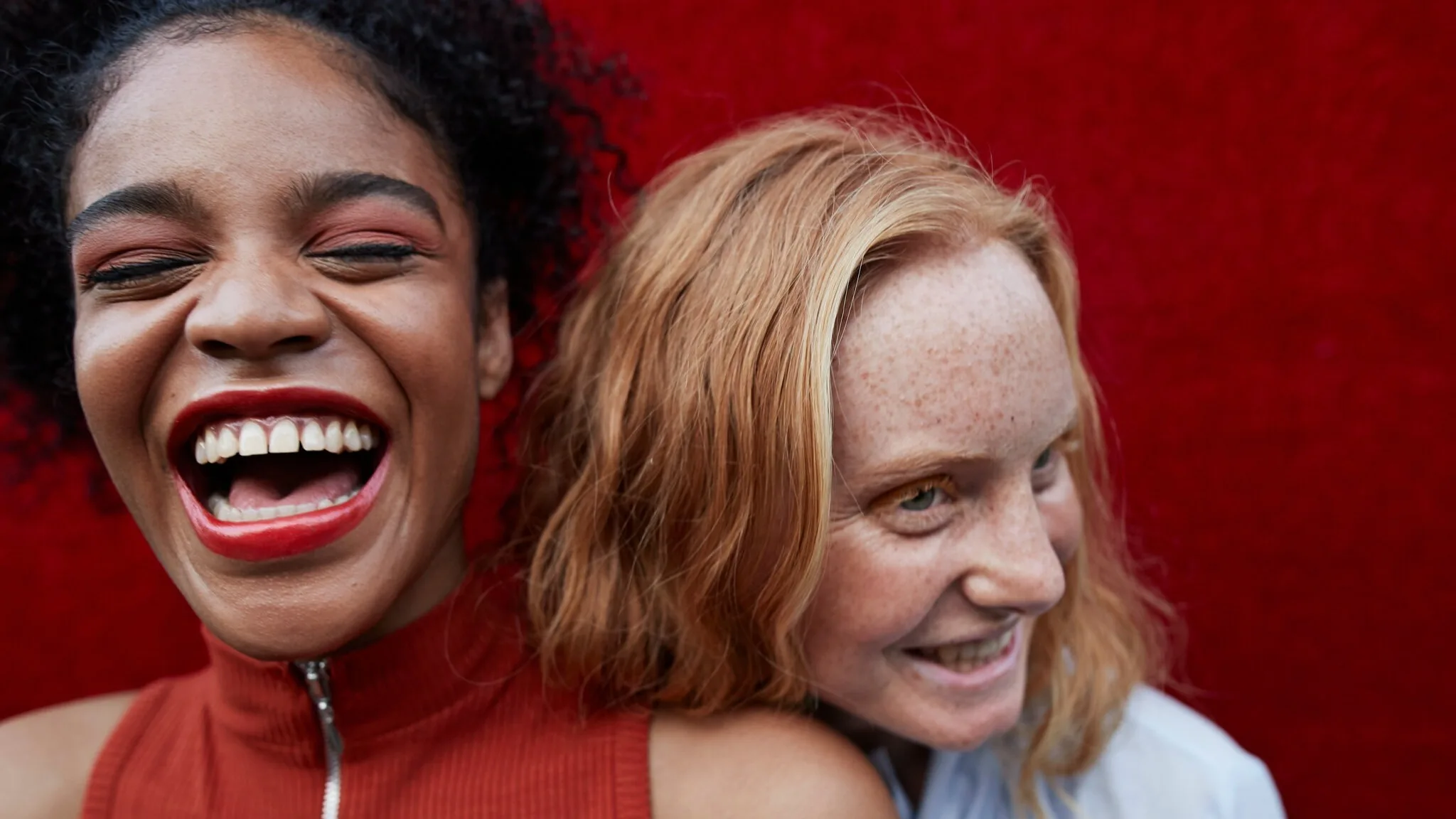 Two women smiling and laughing close together in front of a vibrant red background. One has dark curly hair and wears a sleeveless red top; the other has red hair and freckles, wearing a light blue shirt.