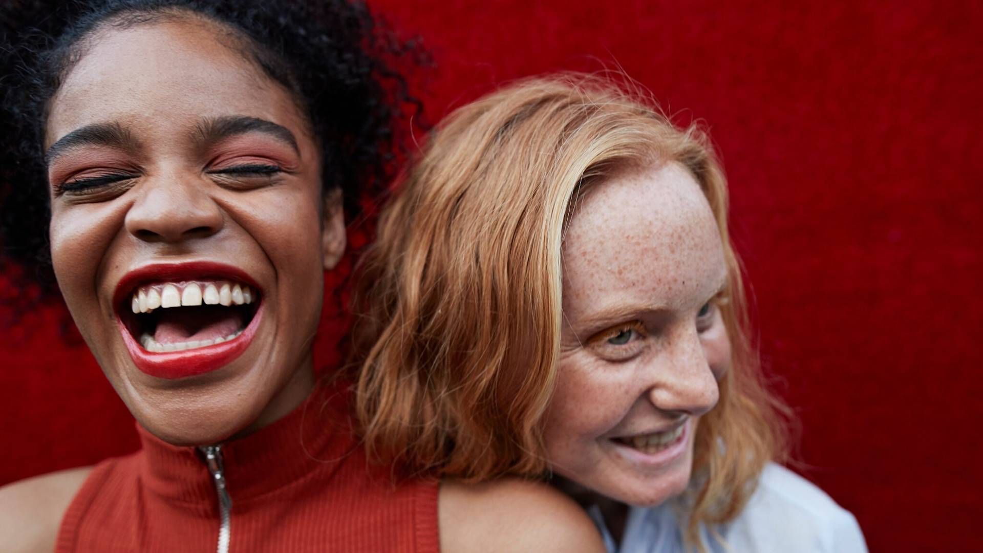 Two women smiling and laughing close together in front of a vibrant red background. One has dark curly hair and wears a sleeveless red top; the other has red hair and freckles, wearing a light blue shirt.