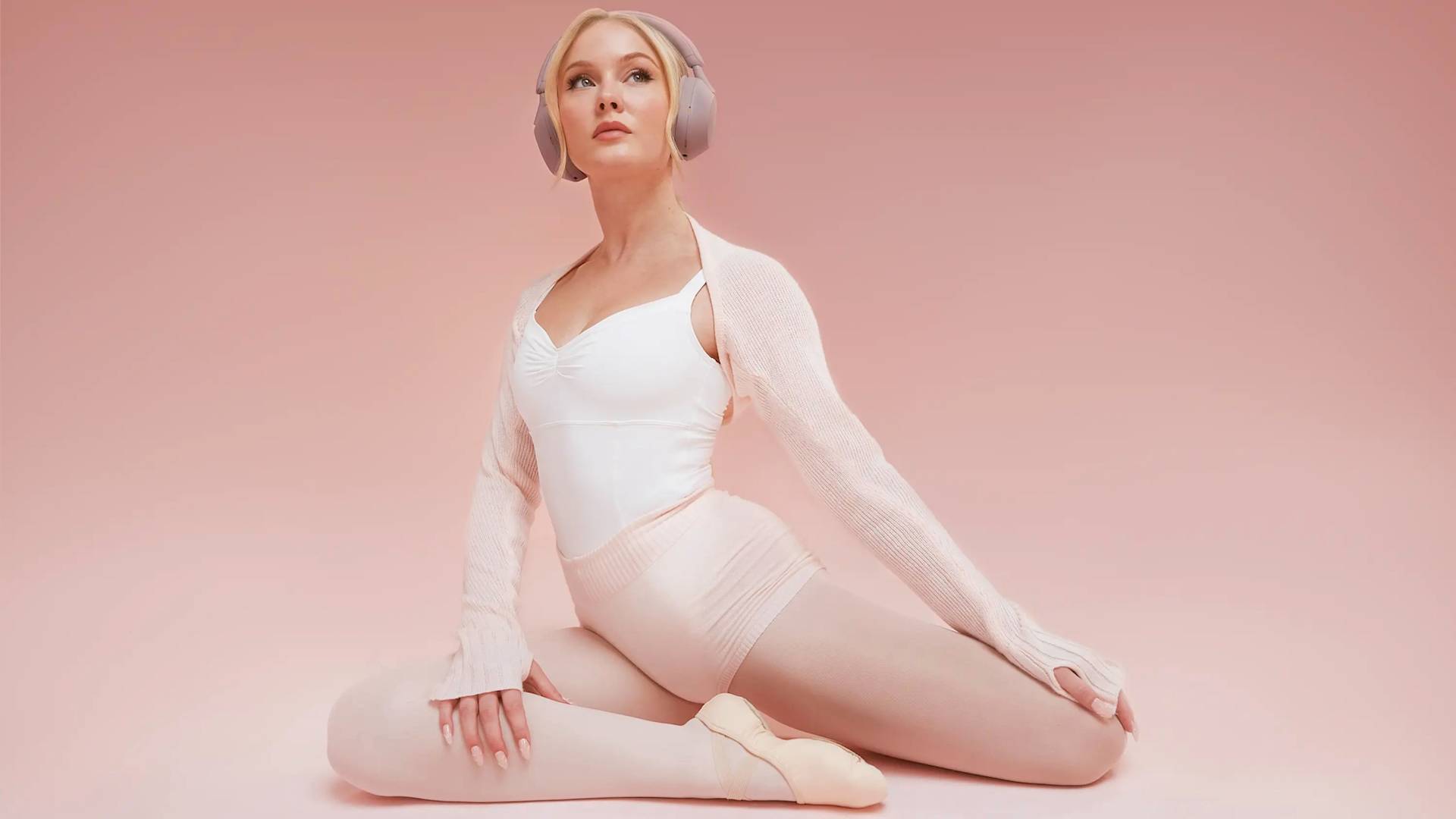 A ballet dancer in a white leotard and tights sits in a stretching pose on a pink background, wearing headphones and a light sweater, looking upwards with a calm expression.