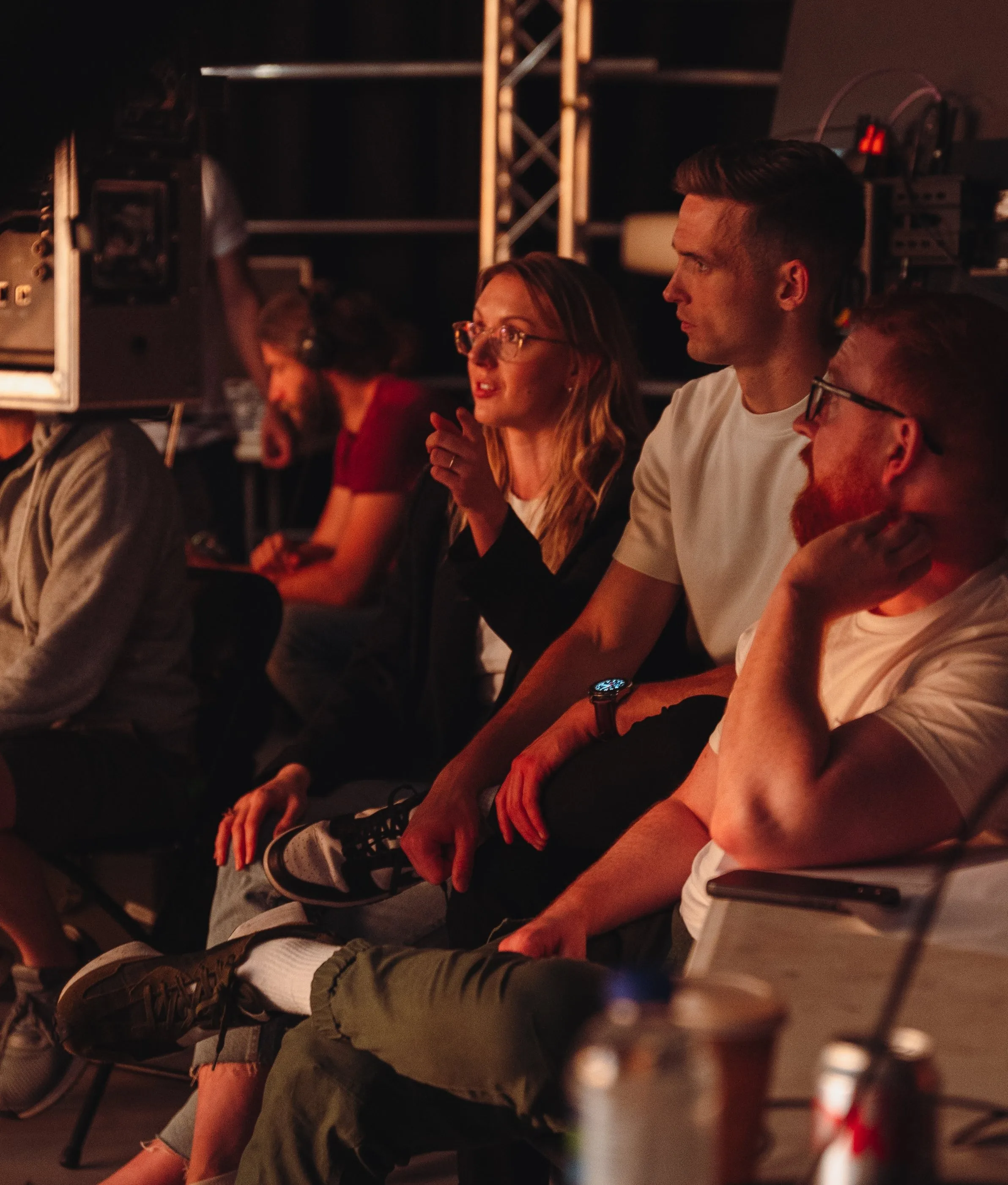 A group of people sit closely together in a dimly lit room, focused intently on something off-camera. One woman gestures while talking, and others listen attentively. Equipment and drinks are visible in the foreground.