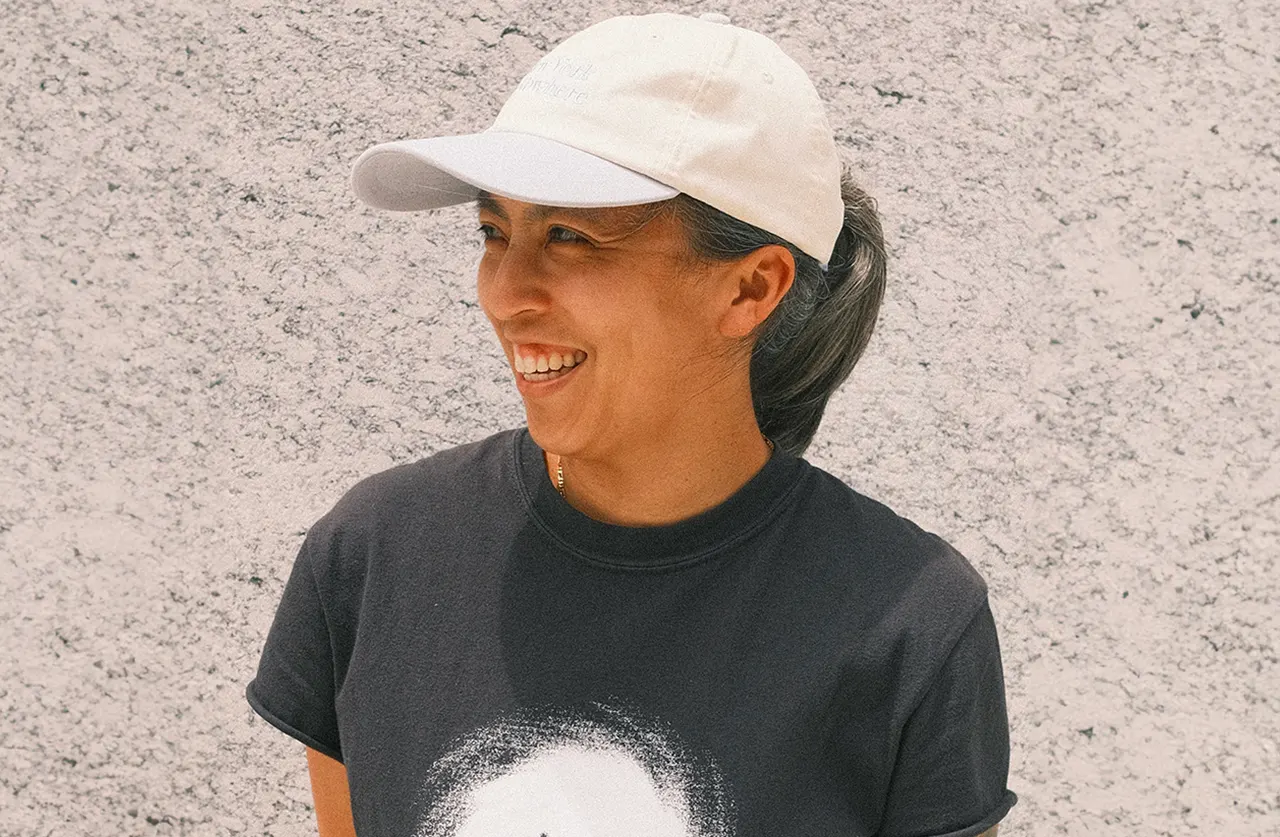 A person wearing a light-colored baseball cap and a black graphic t-shirt smiles while standing in front of a textured, light-colored wall.