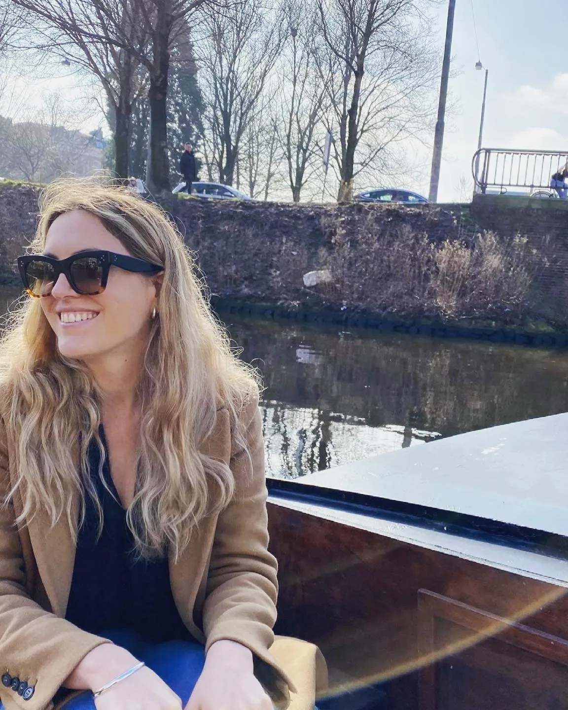 A woman with long blonde hair and sunglasses smiles while sitting on a boat by a calm canal, with bare trees and a stone wall in the background on a sunny day.