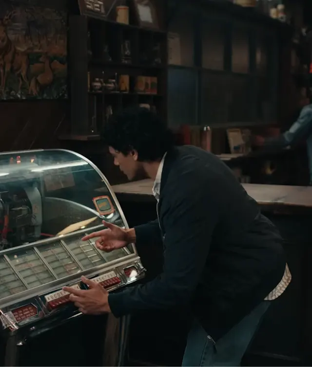 A person in a dark jacket is selecting a song on a vintage jukebox in a dimly lit room with shelves and various items in the background.