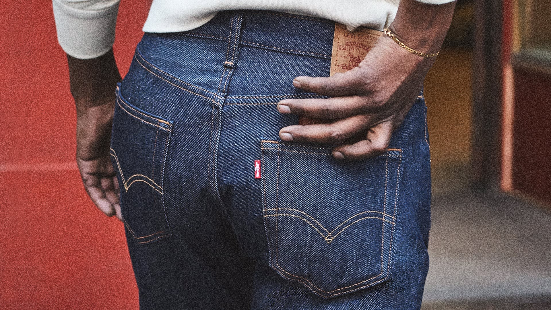 A person wearing blue denim jeans with one hand resting on the back pocket, showing a brown label and a small red tag on the pocket. The background features a red wall.