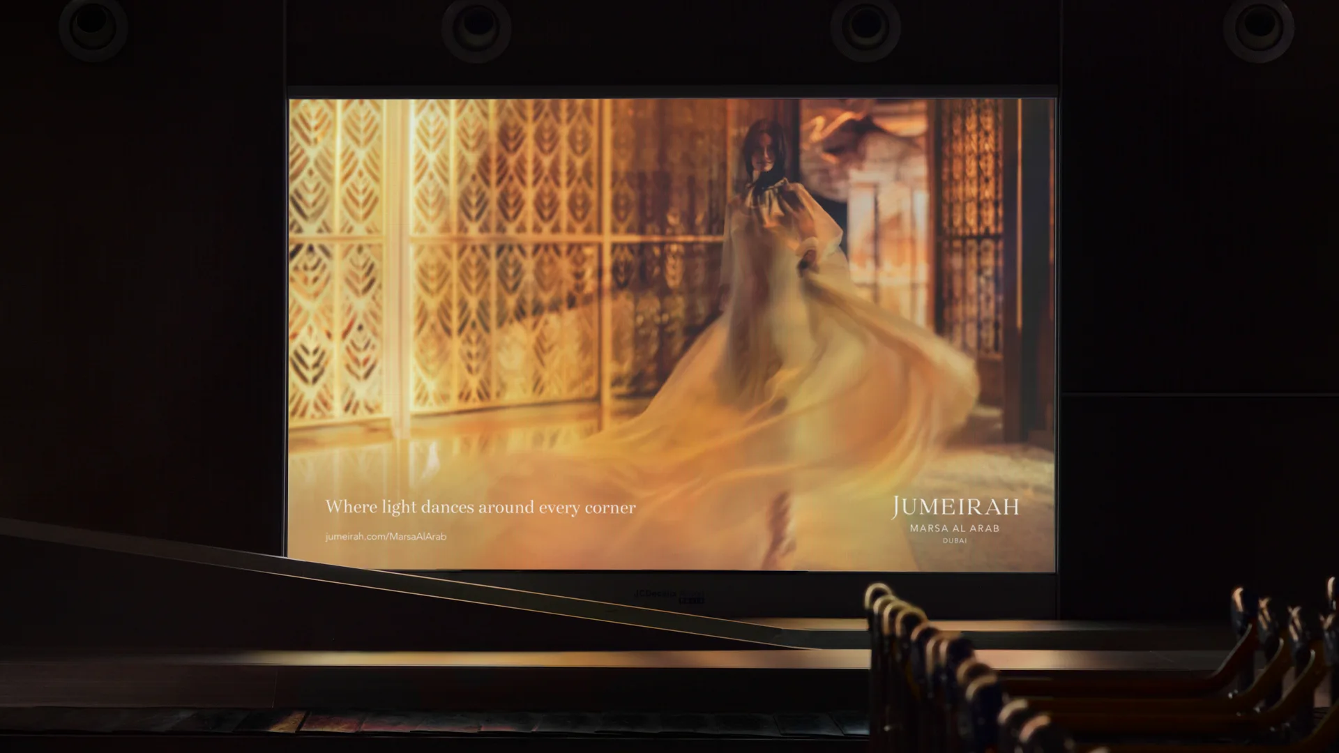 A projected advertisement shows a woman in a flowing gown spinning gracefully in a golden, ornate room. The text reads: Where light dances around every corner. Jumeirah Marsa Al Arab Dubai.