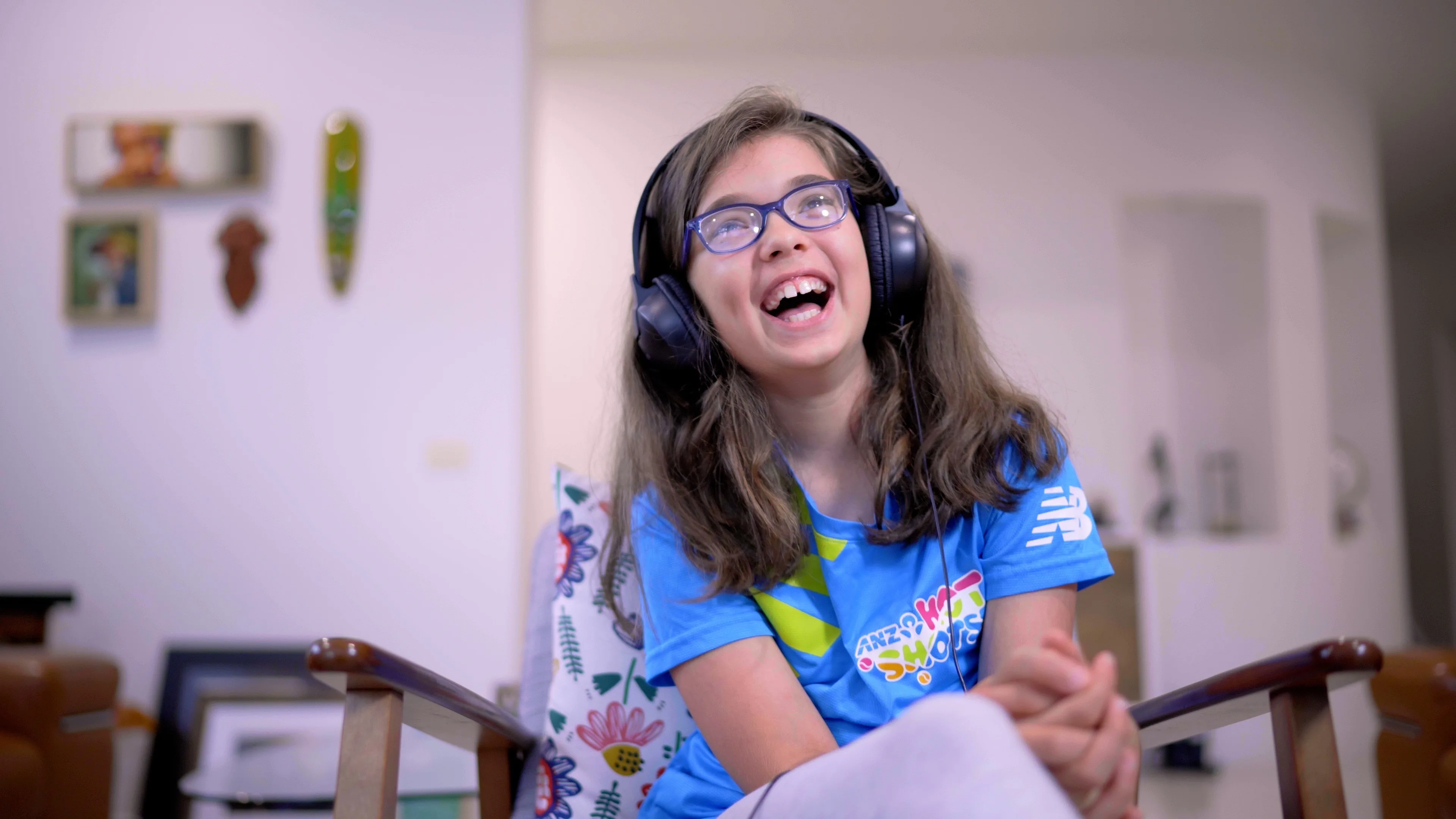 A young girl with glasses, wearing headphones and a blue t-shirt, sits on a chair indoors, smiling and laughing joyfully.