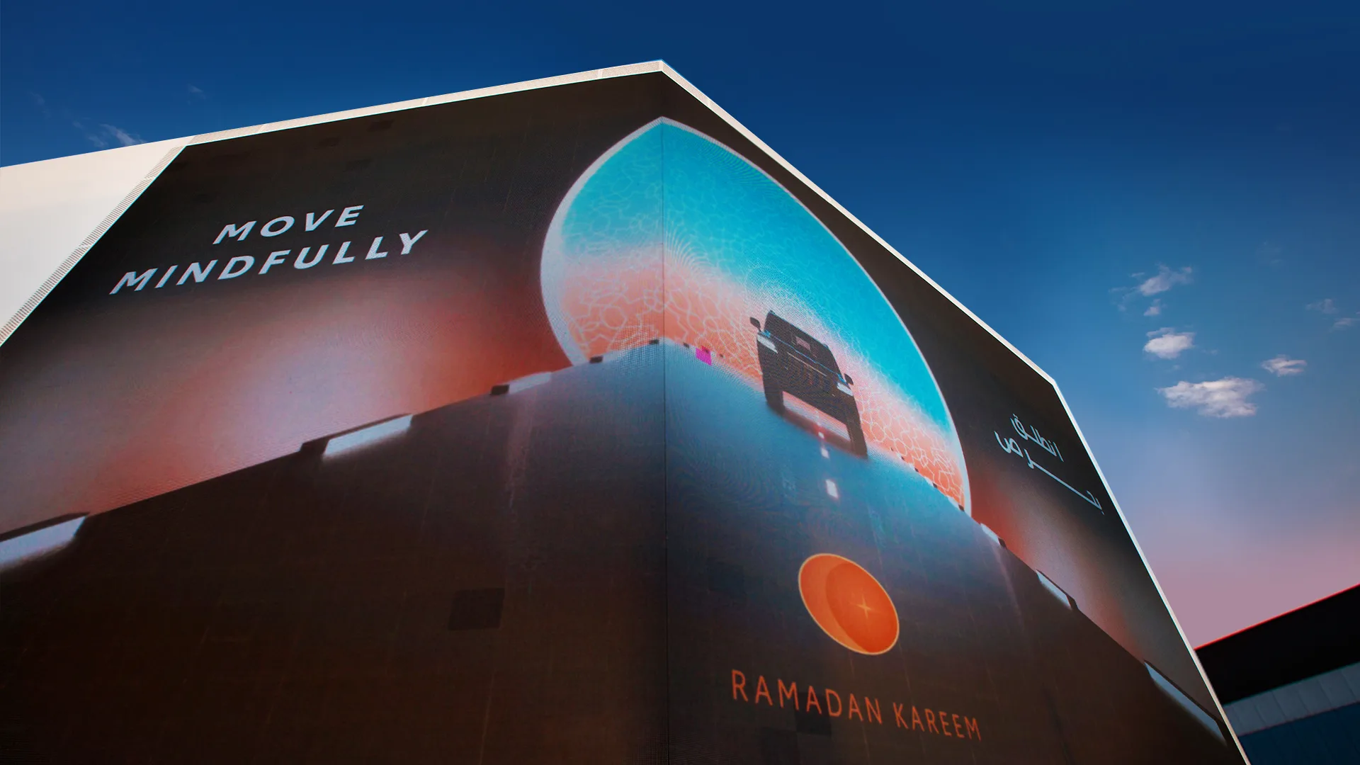 A large building billboard displays the message MOVE MINDFULLY and RAMADAN KAREEM with an abstract design featuring a glowing lantern against a blue and orange gradient background, under a clear sky.