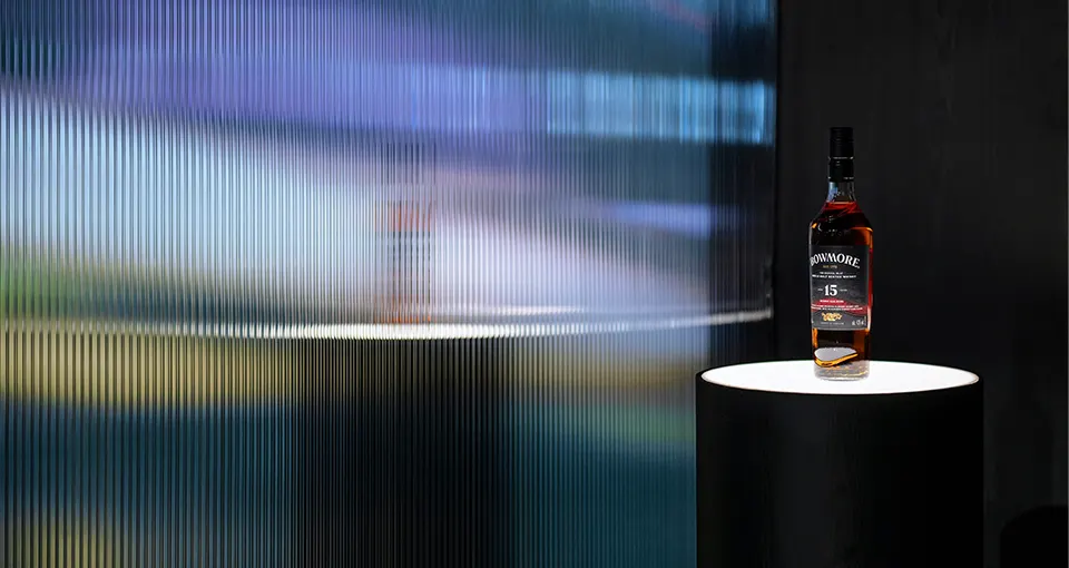 A bottle of Glenmorangie 15-year-old Scotch whisky stands on a round black pedestal, spotlighted against a blurred, colorful, and textured glass background.