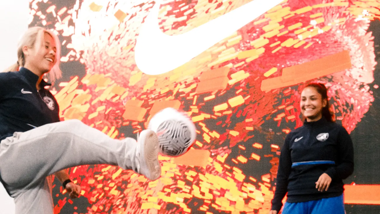 Two women in sportswear smile as one of them kicks a soccer ball. A large, vivid red and orange Nike logo is displayed on the screen in the background.