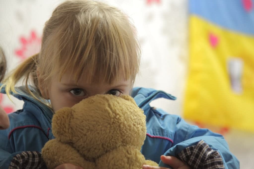 A young child in a blue jacket holds a brown teddy bear up to their face, peeking over it with wide eyes. The background is softly blurred with hints of flowers and colorful shapes.