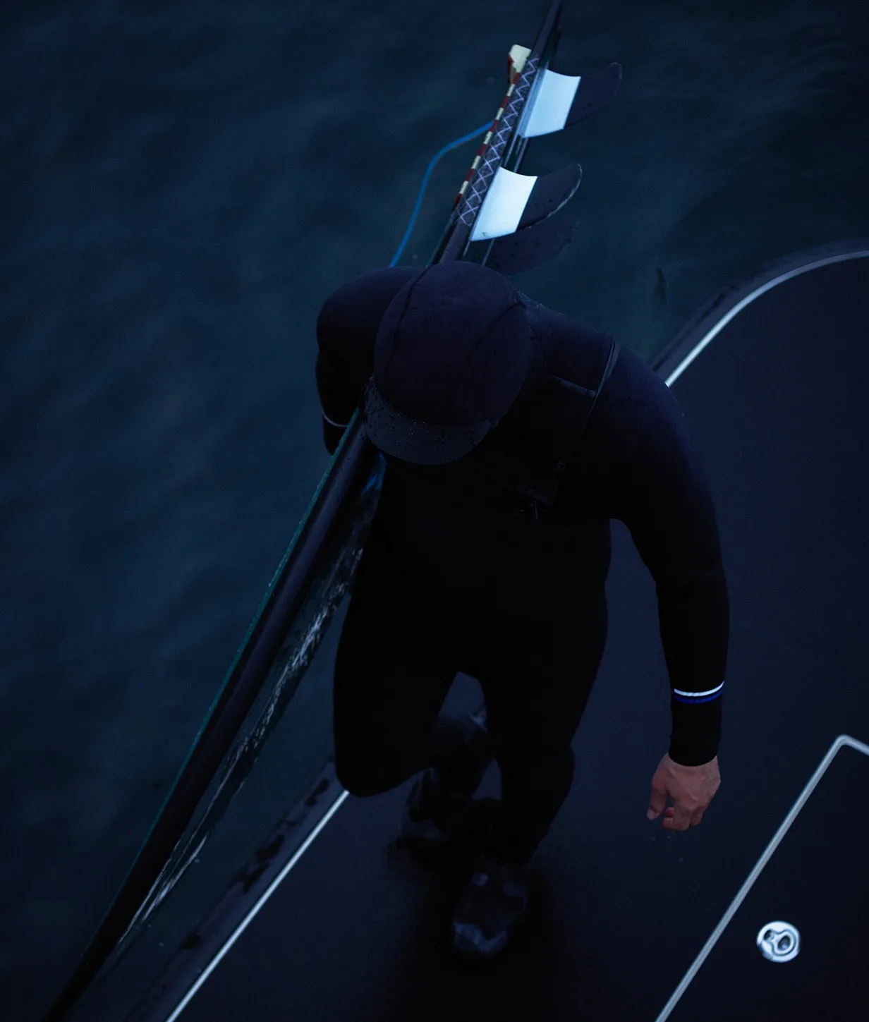 A person in a black wetsuit and cap stands on a platform above dark water, holding a surfboard with three fins under their arm. Their face is not visible as the image is shot from above.