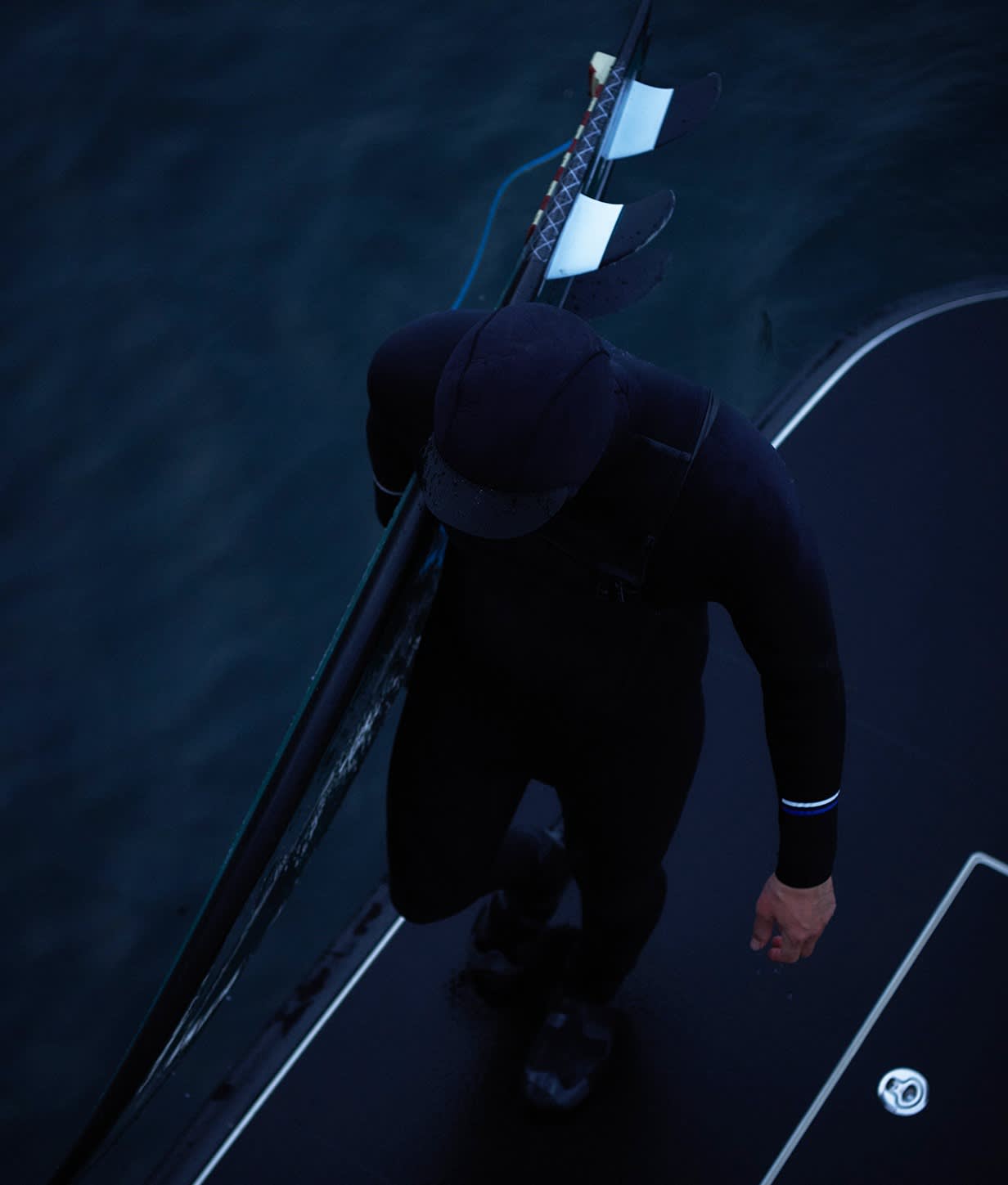 A person in a black wetsuit and cap stands on a platform above dark water, holding a surfboard with three fins under their arm. Their face is not visible as the image is shot from above.
