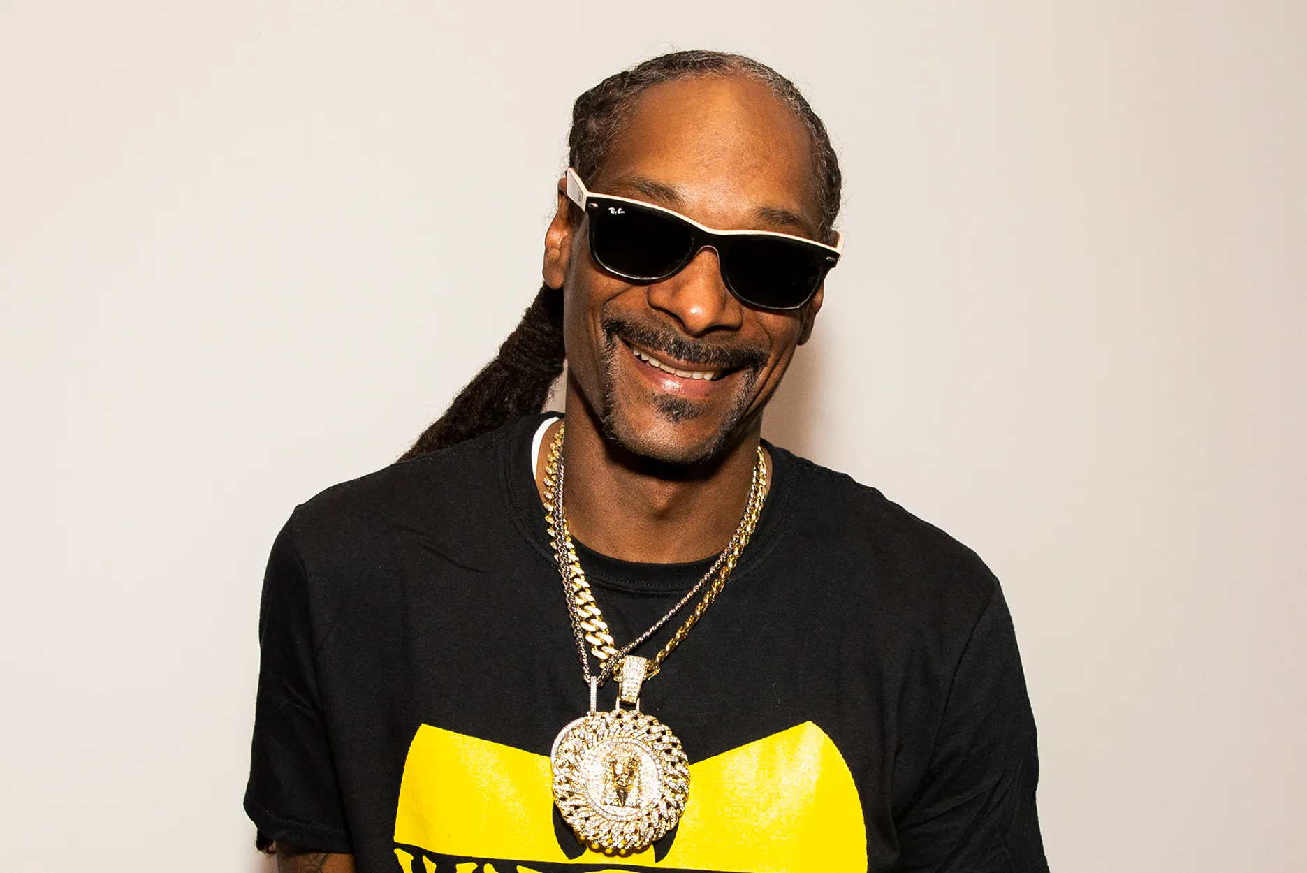A man with long braided hair wears black sunglasses, a black and yellow t-shirt, and large gold chains with pendants. He is smiling and standing against a plain light-colored background.