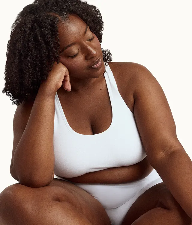A woman with curly hair sits in a relaxed pose, resting her head on her hand and wearing a white sports bra and underwear against a plain background.