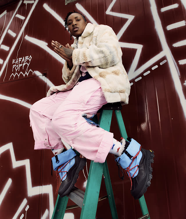 A person wearing a furry jacket, pink pants, and chunky blue and black boots sits on a green ladder against a red wall with white graffiti patterns. They have their hands together and look confidently at the camera.