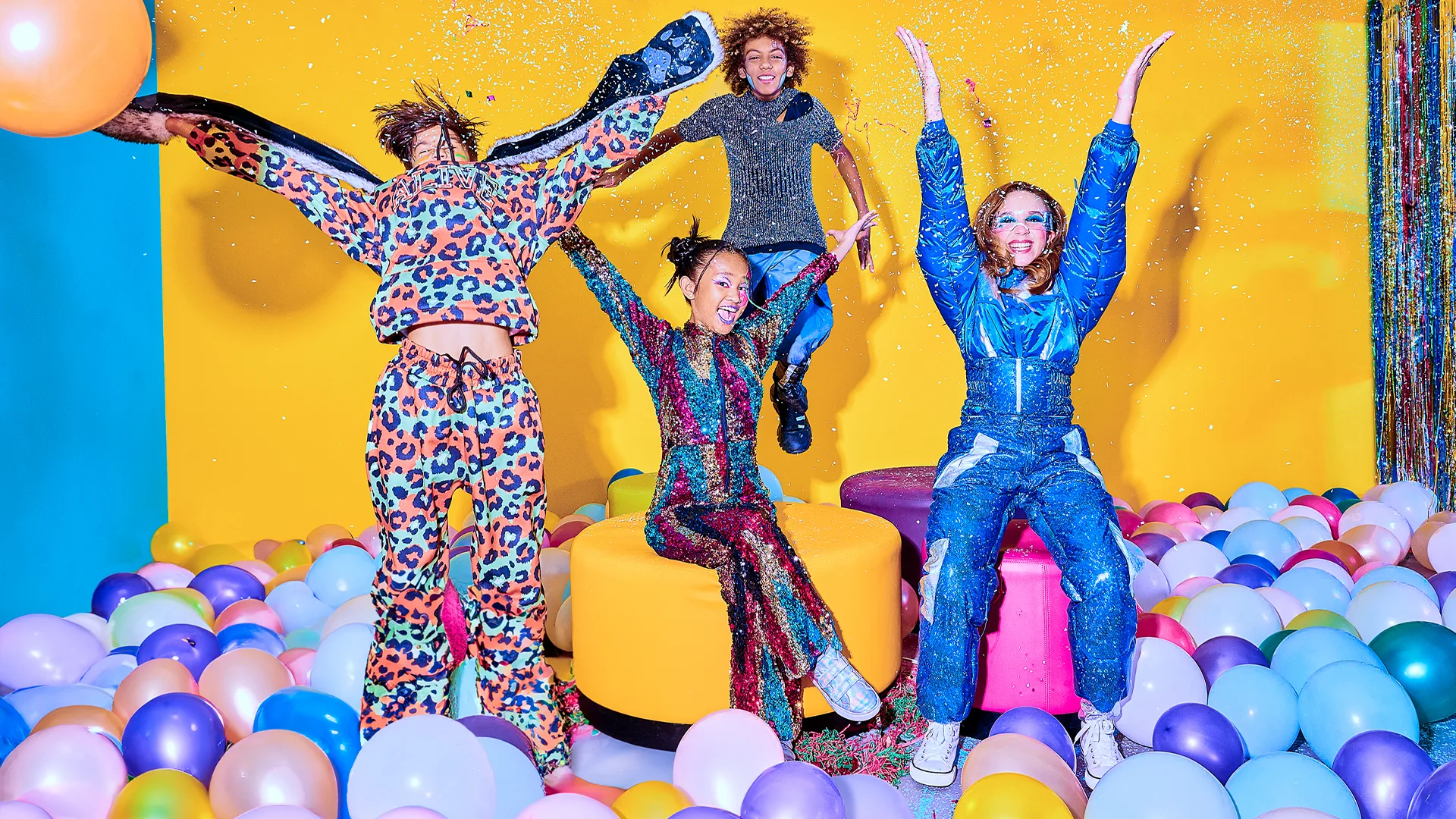 Four people in colorful, sparkly outfits celebrate in a room filled with pastel balloons against a yellow background. Two stand with arms raised, one sits smiling, and one jumps in the air, all surrounded by confetti.