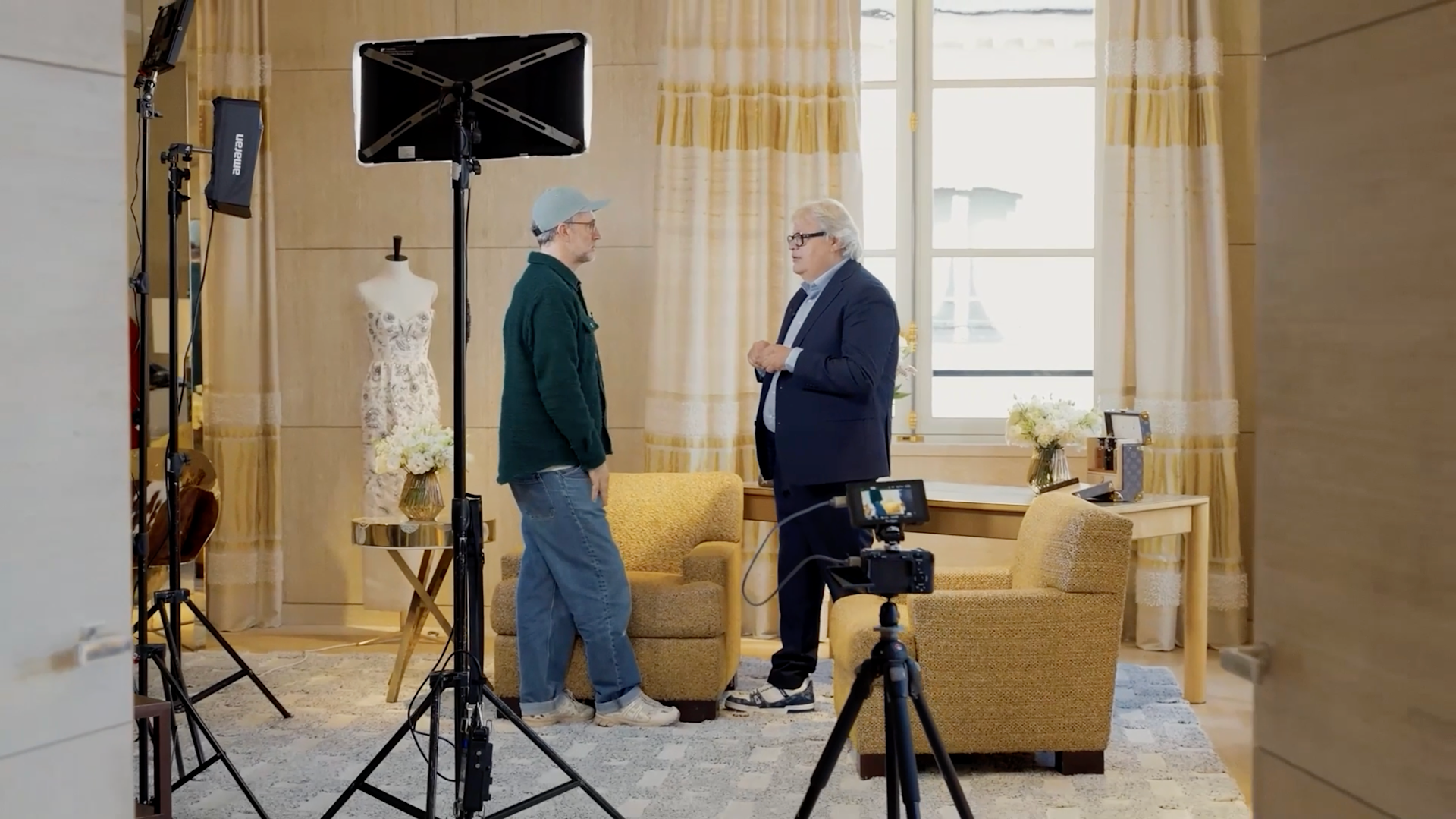 Two men stand and talk in a well-lit, elegant room set up for filming, with a video camera, tripod, and lights. Behind them are yellow armchairs, a mannequin in a dress, and large windows with curtains.