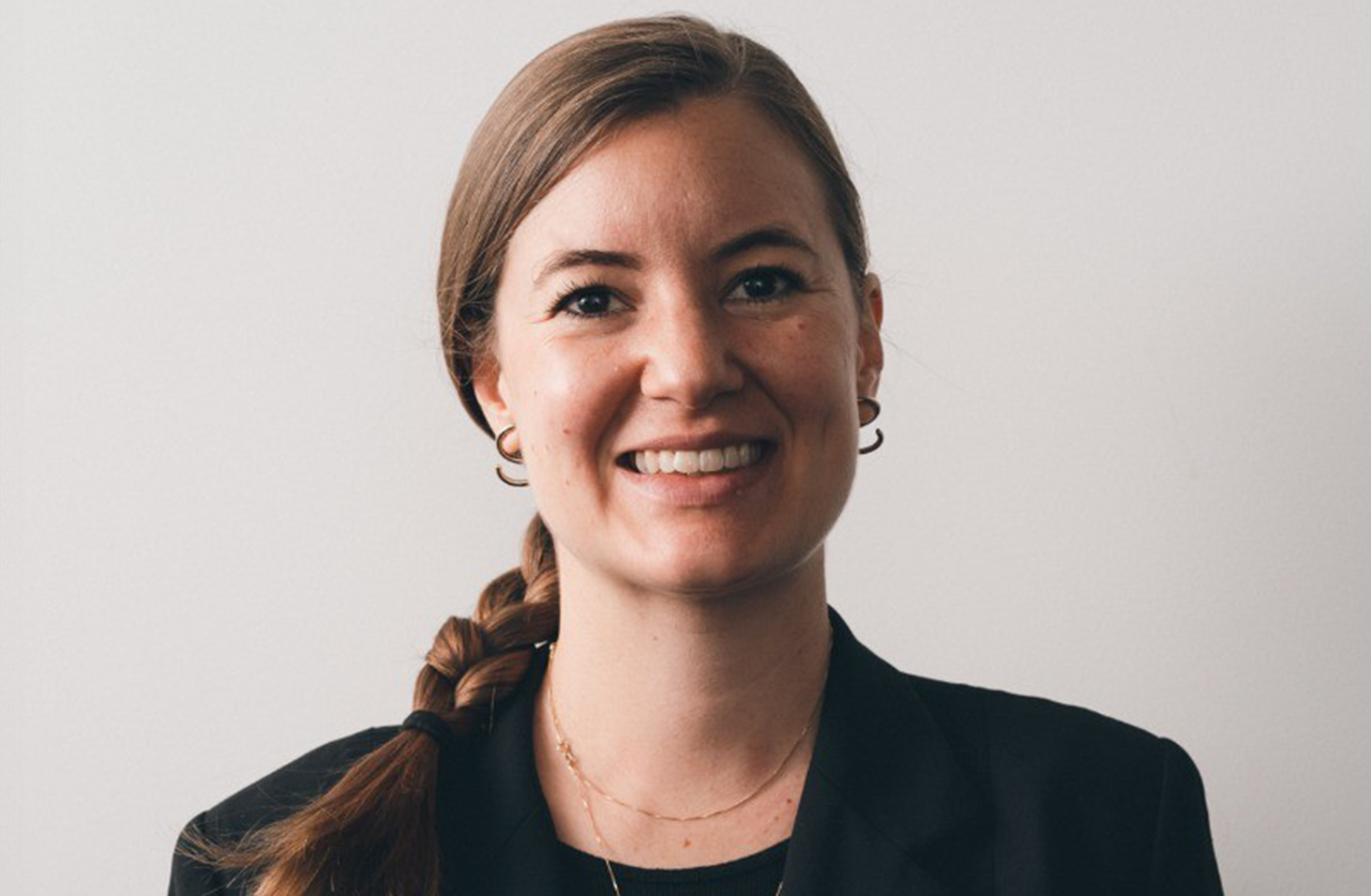A woman with light brown hair in a braid, wearing a black blazer, double hoop earrings, and a necklace, smiles at the camera against a plain, light background.
