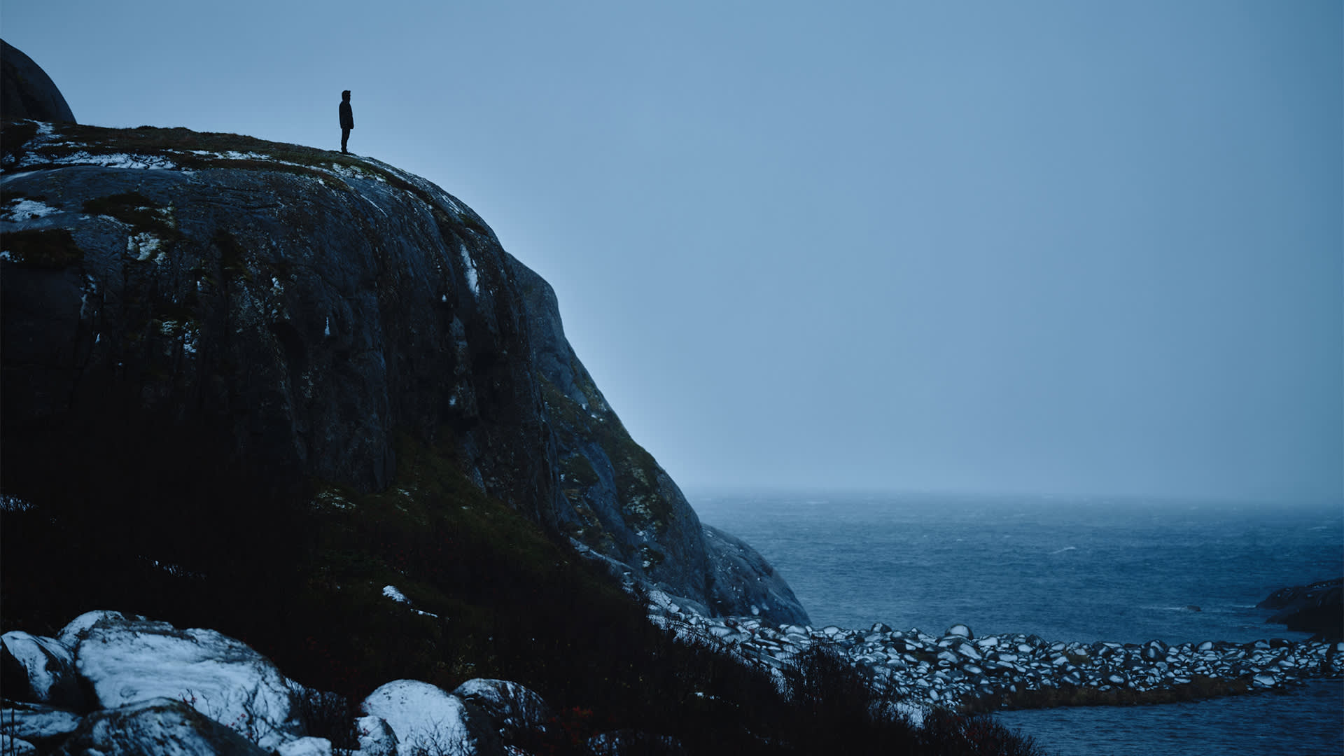 A solitary figure stands on top of a rocky cliff overlooking a vast, misty ocean under an overcast sky, creating a moody, atmospheric scene.