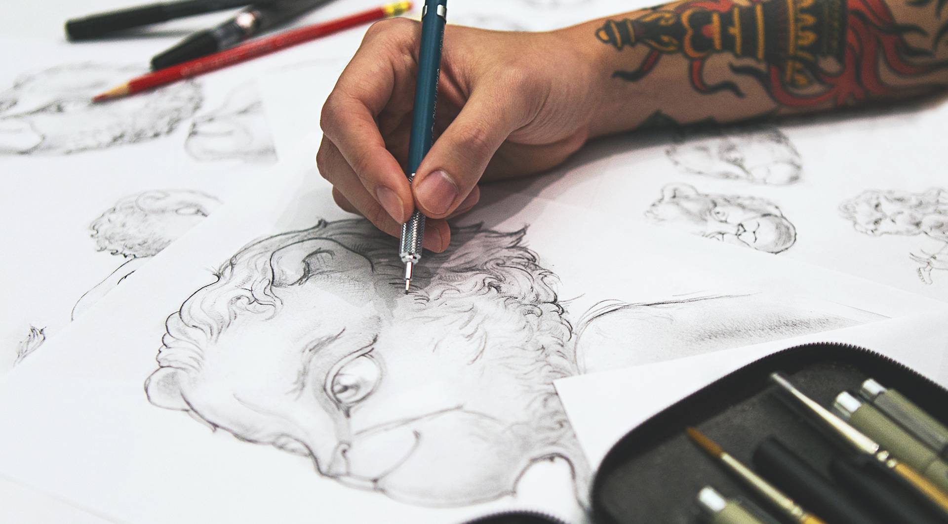 A tattooed arm holds a pen drawing the Future Lions character, a smiling lion with big eyes. Underneath the drawing lies sketches of different angles of the lion, in the bottom right-hand corner is a pencil case with paintbrushes and pens.