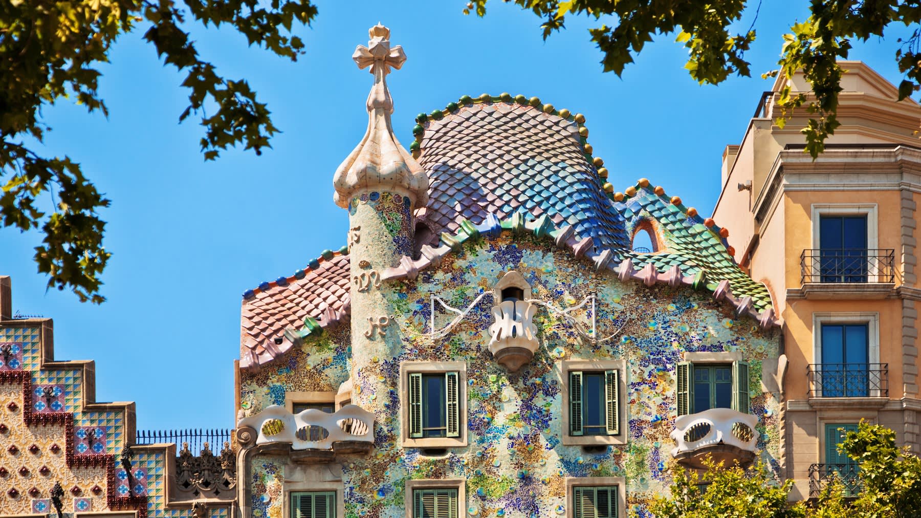 Colorful mosaic façade and whimsical, curved roof of Casa Batlló in Barcelona, featuring a cross-topped turret and ornate balconies, framed by leafy tree branches under a clear blue sky.