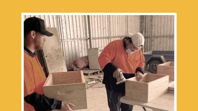 Two people wearing orange shirts work on wooden boxes in a workshop; one sands a box while the other holds another box. Both are wearing caps, and the background shows tools and workshop materials.