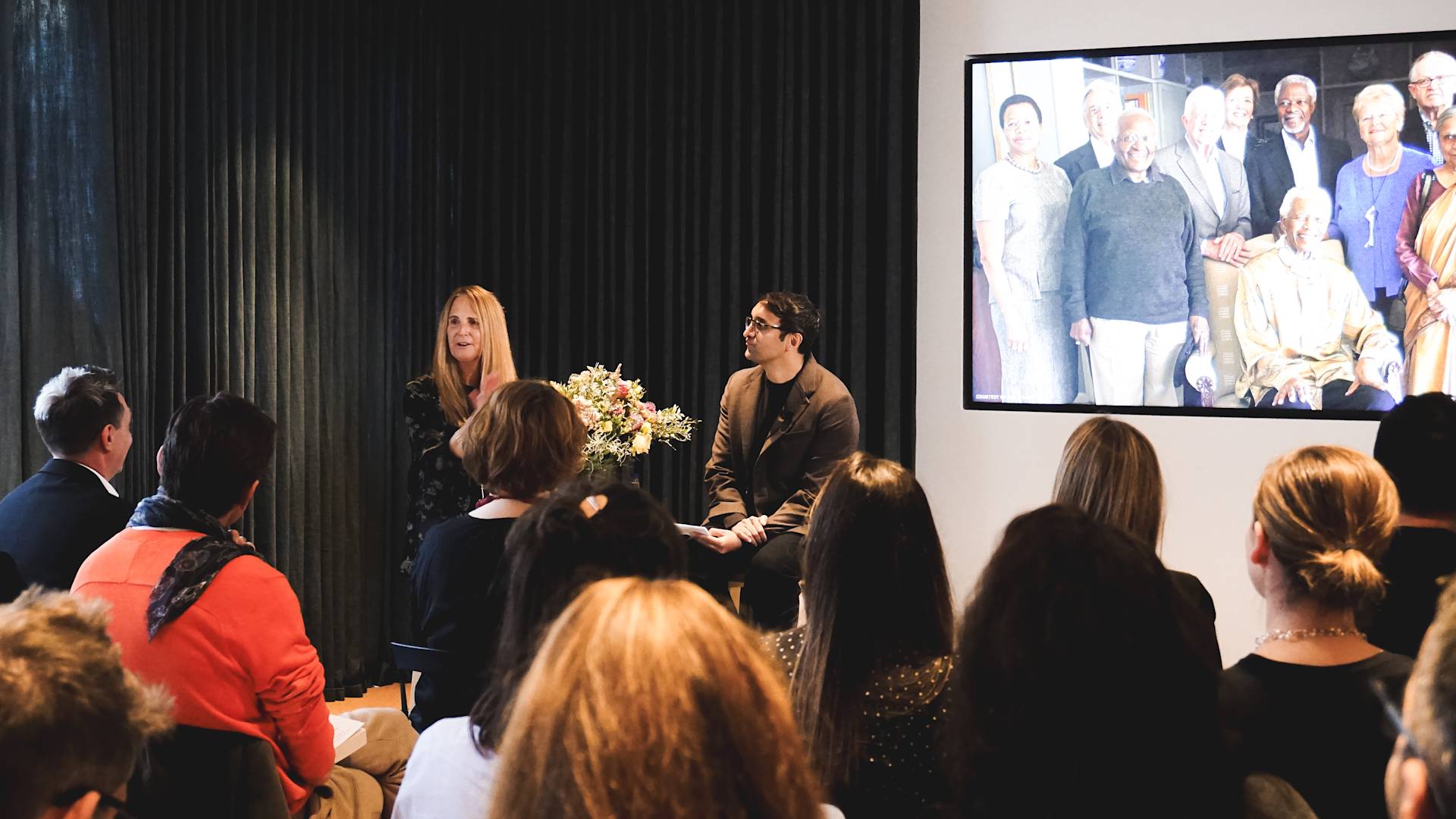 A woman and a man sit and speak in front of an audience in a room. Behind them is a large screen displaying a group photo of older adults. The audience listens attentively.