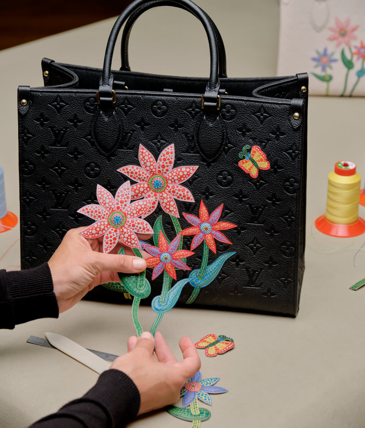 A person arranges colorful embroidered flower patches in front of a black Louis Vuitton handbag, decorated with a butterfly patch. Spools of thread and tools are visible on the beige tabletop.