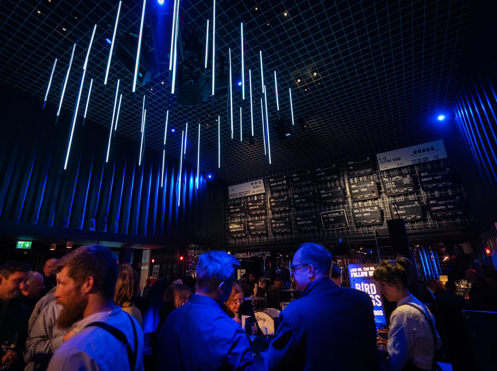 People gather in a dimly lit, modern bar or event space with blue LED lights hanging from the ceiling and an illuminated drinks menu on the back wall. The atmosphere is lively and social.