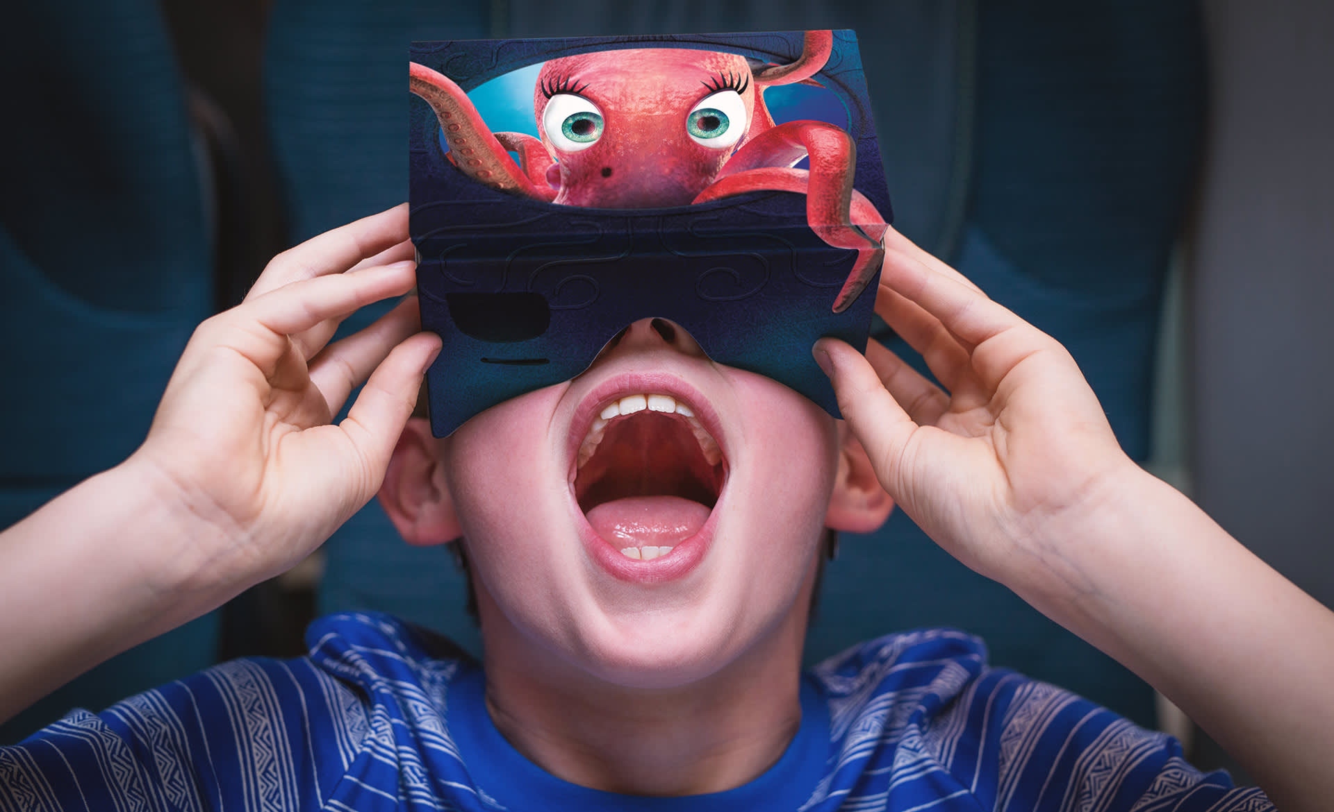 A child wearing a blue striped shirt holds a cardboard VR headset with a cartoon octopus design over their eyes, mouth open wide in excitement or surprise.