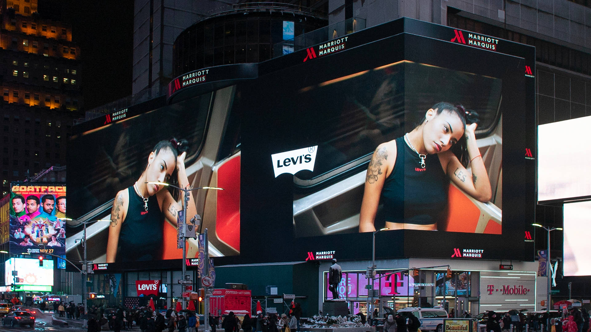 Large digital billboards on a city building display a Levi’s advertisement featuring a woman with tattoos leaning her head to one side. Neon lights and store signs, including T-Mobile, are visible below in the busy city scene.