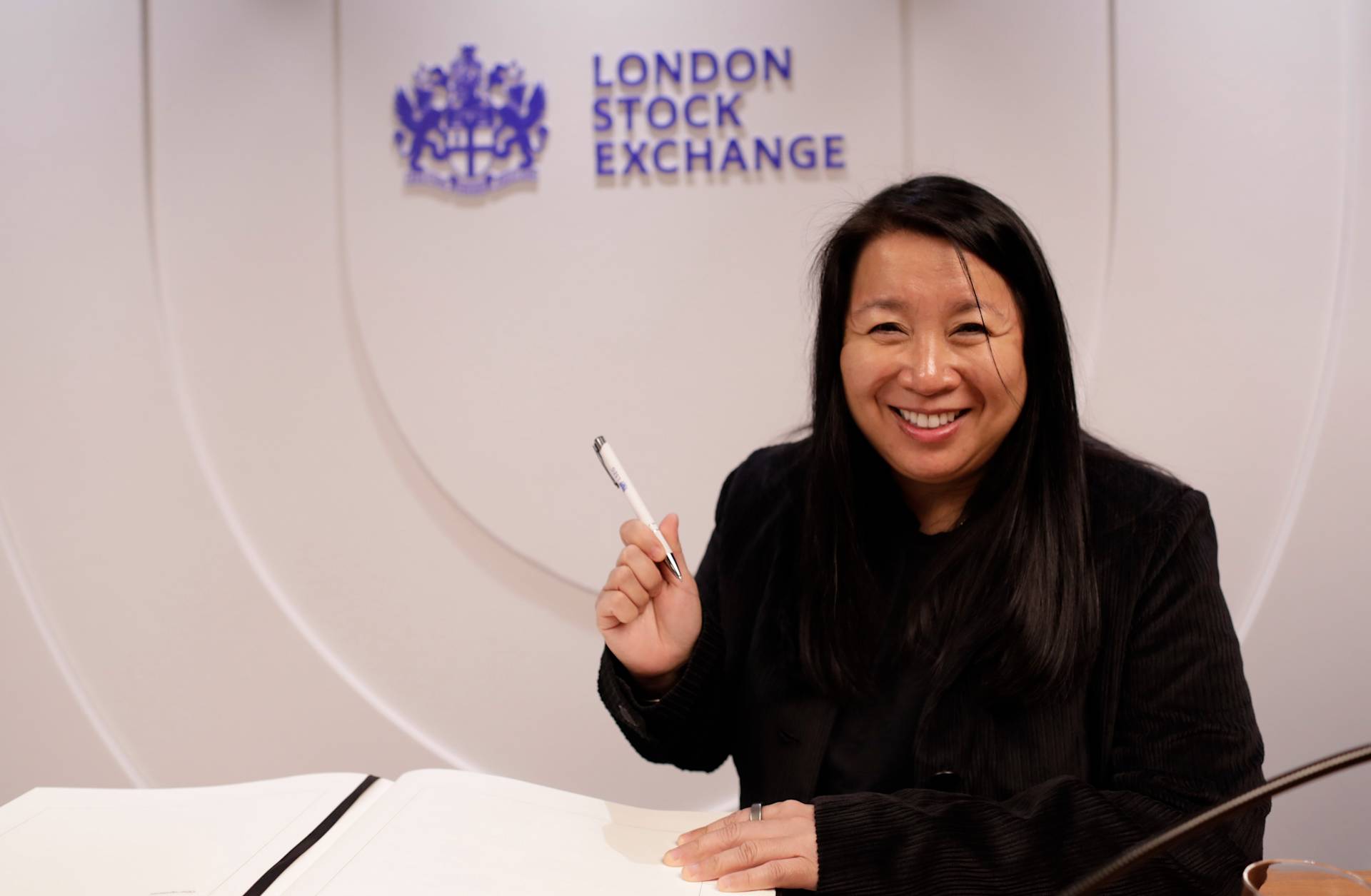 A smiling woman holds a pen and prepares to sign a book at a table, with the London Stock Exchange logo and name displayed on the wall behind her.