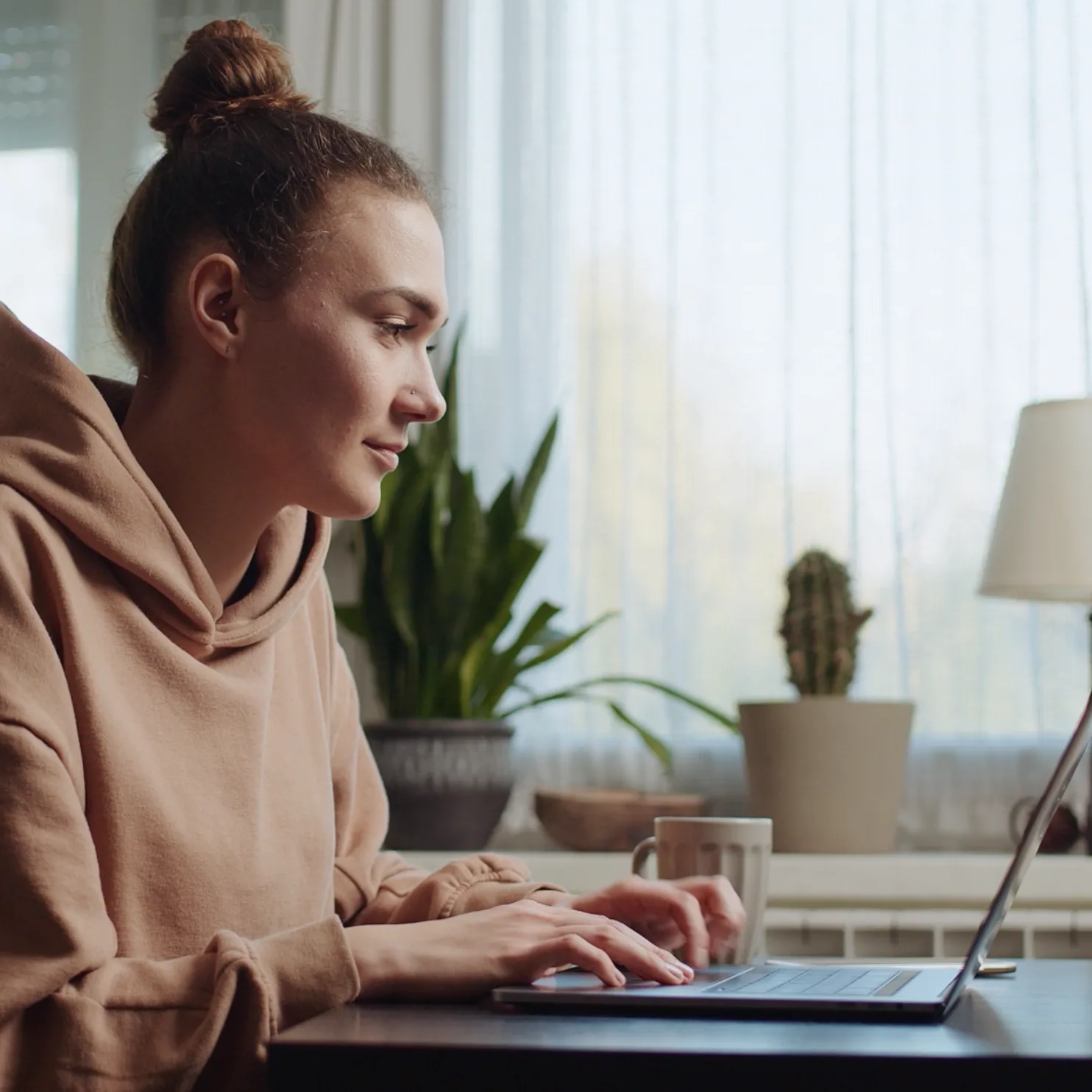 A person wearing a tan hoodie sits at a table using a laptop, with a mug beside them. In the background, potted plants and a lamp are next to a window with sheer curtains.