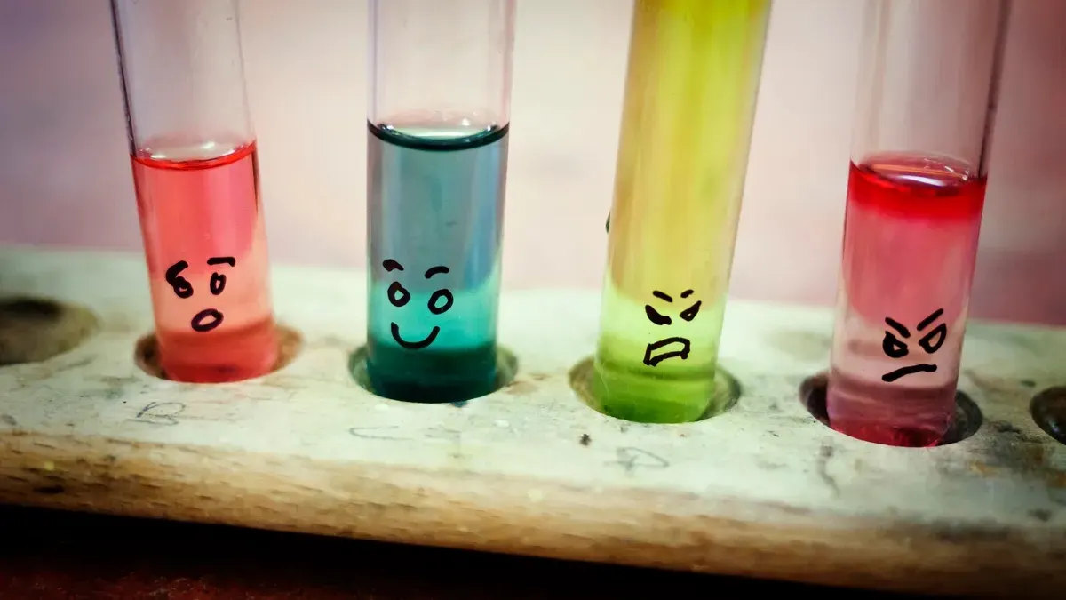 Four test tubes with different colored liquids (pink, blue, yellow, and red) sit in a wooden holder. Each test tube has a unique drawn face expressing various emotions like surprise, happiness, anger, and annoyance.