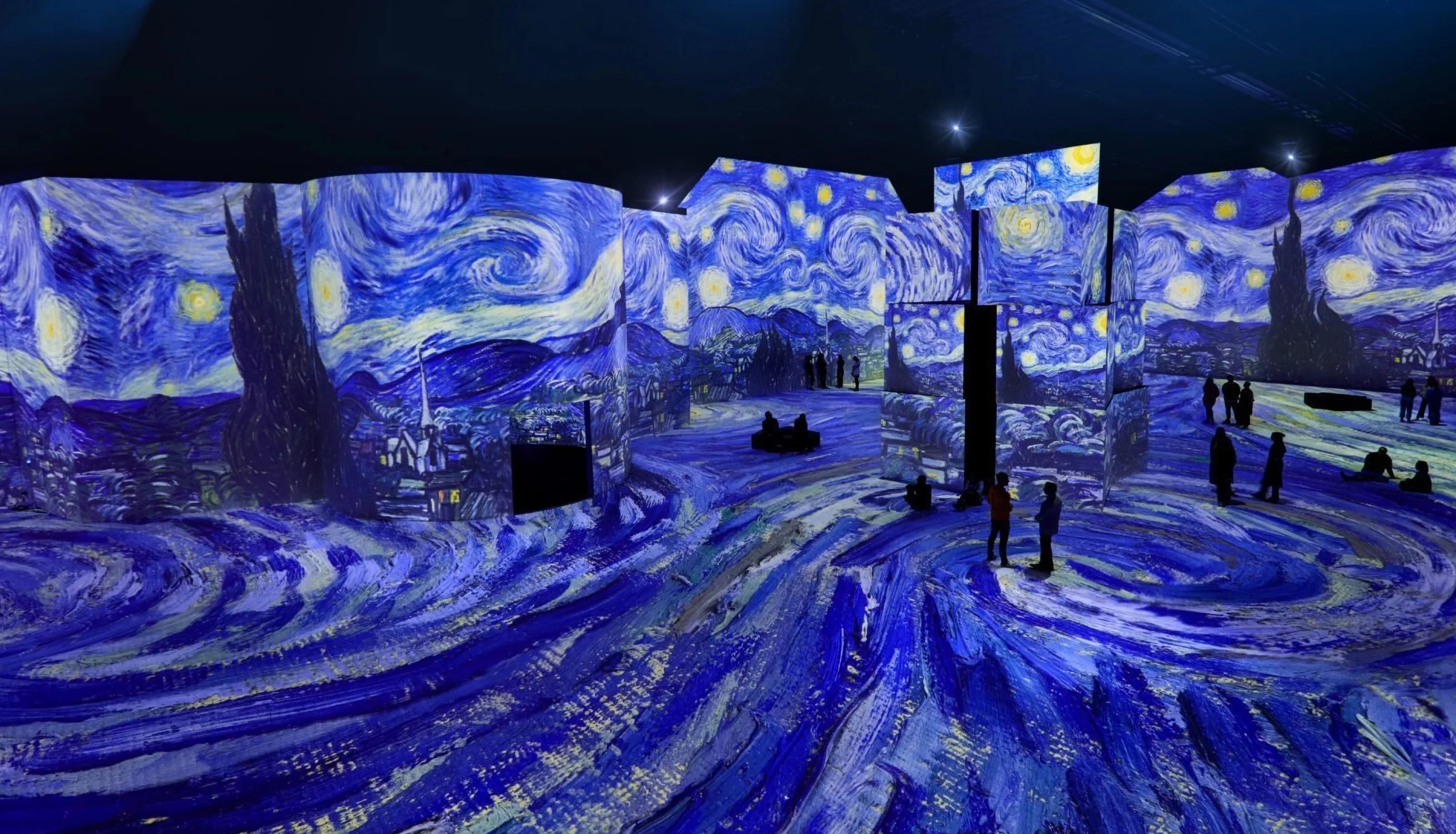 An immersive art exhibit features walls and floors illuminated with swirling blue and yellow patterns inspired by Van Goghs Starry Night, with visitors silhouetted as they explore the vibrant, immersive space.