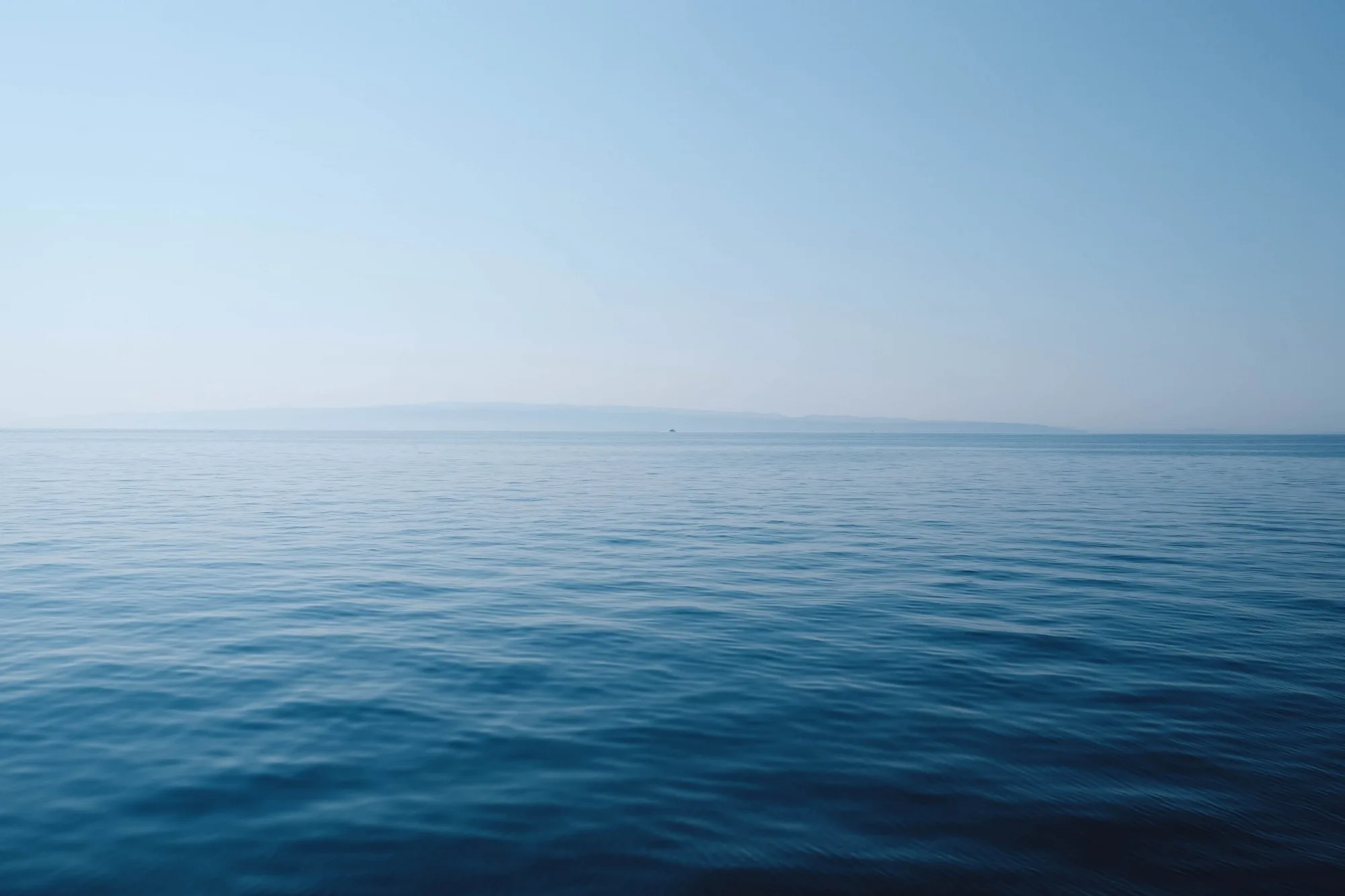 Calm, blue ocean water stretches out to the horizon under a clear sky, with faint, distant land visible on the horizon. The scene is tranquil and serene, with gentle ripples on the sea’s surface.