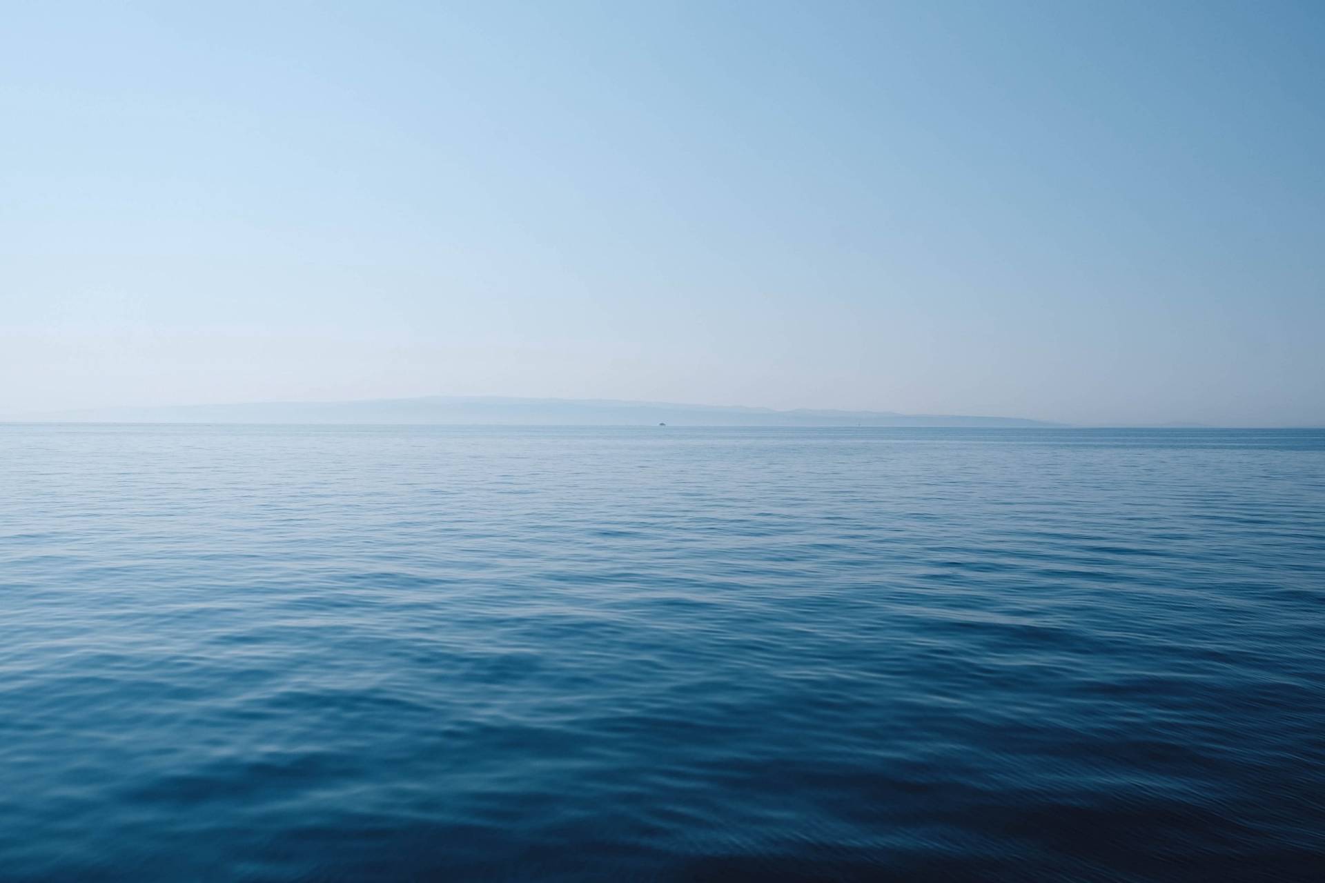 Calm, blue ocean water stretches out to the horizon under a clear sky, with faint, distant land visible on the horizon. The scene is tranquil and serene, with gentle ripples on the sea’s surface.