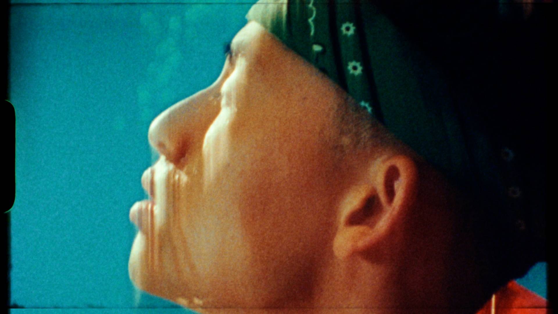 A close-up side profile of a person wearing a green bandana, looking upward against a blue background. Warm light highlights their face, casting gentle shadows and creating a soft, contemplative mood.