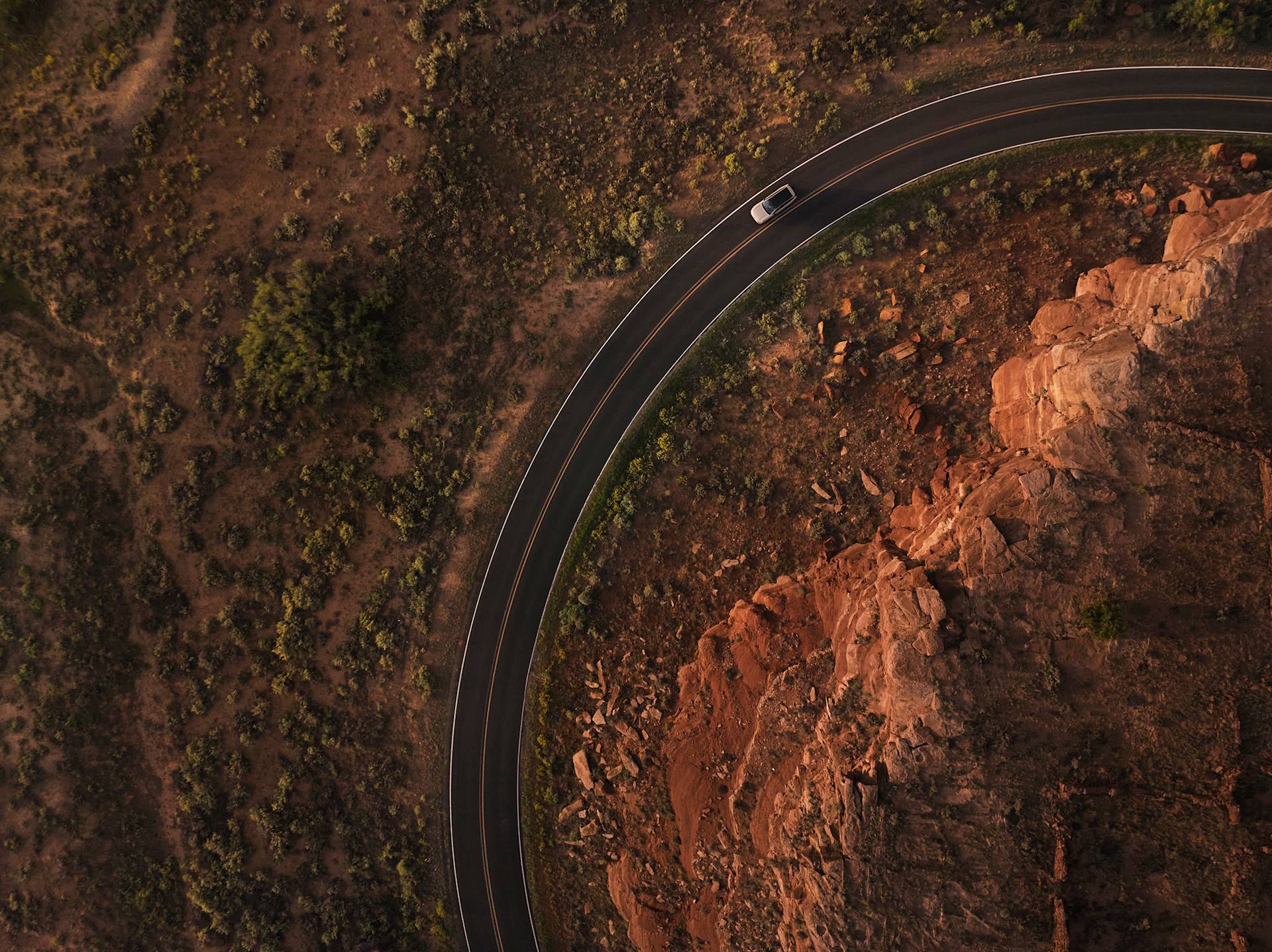 Aerial view of a car driving along a curved road bordering rocky, reddish terrain and sparse vegetation in a desert landscape.