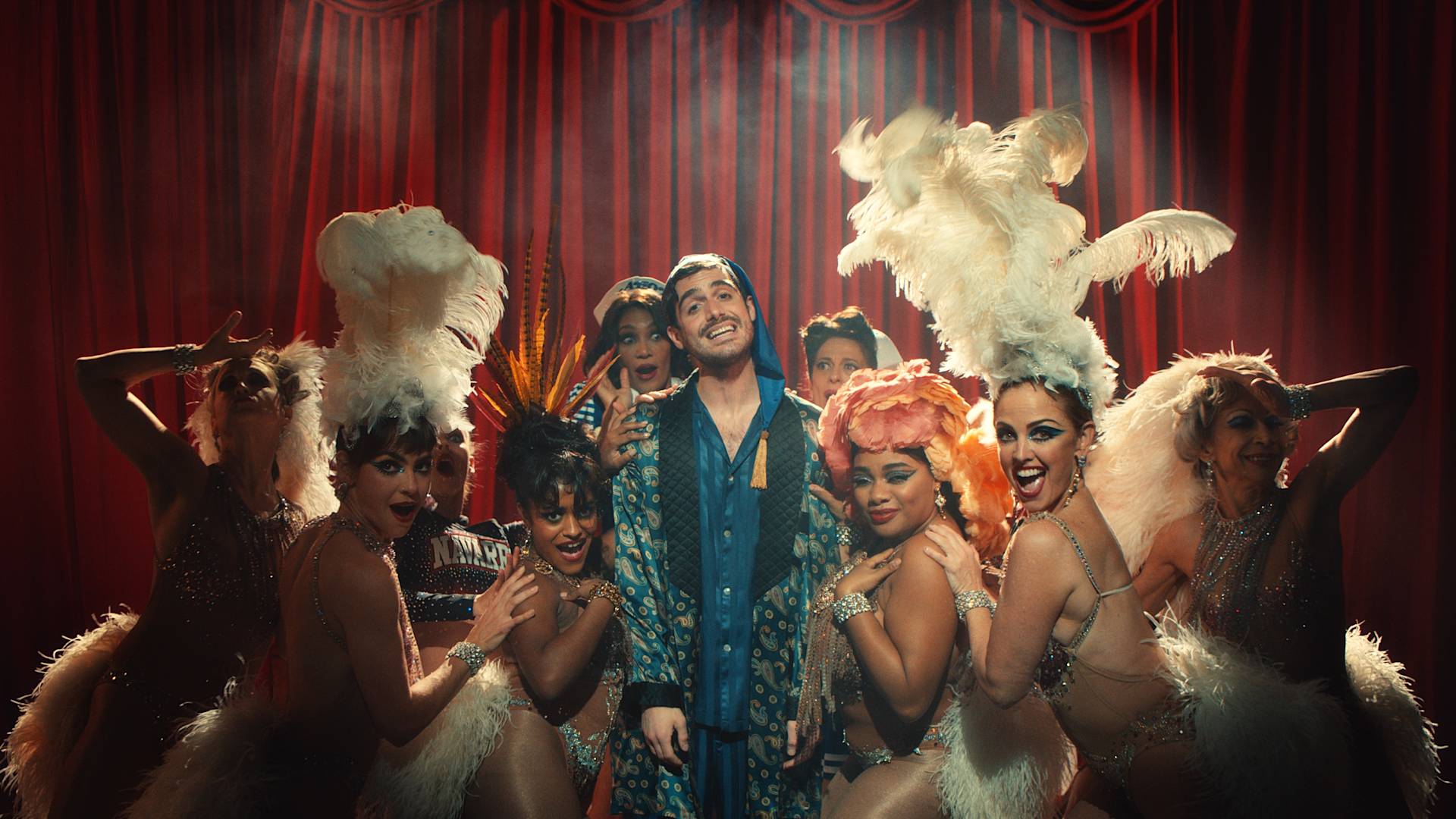 A smiling man in a patterned robe stands center stage, surrounded by glamorous showgirls in feathered headpieces and sparkling costumes, with a red curtain backdrop. The scene is vibrant and theatrical.