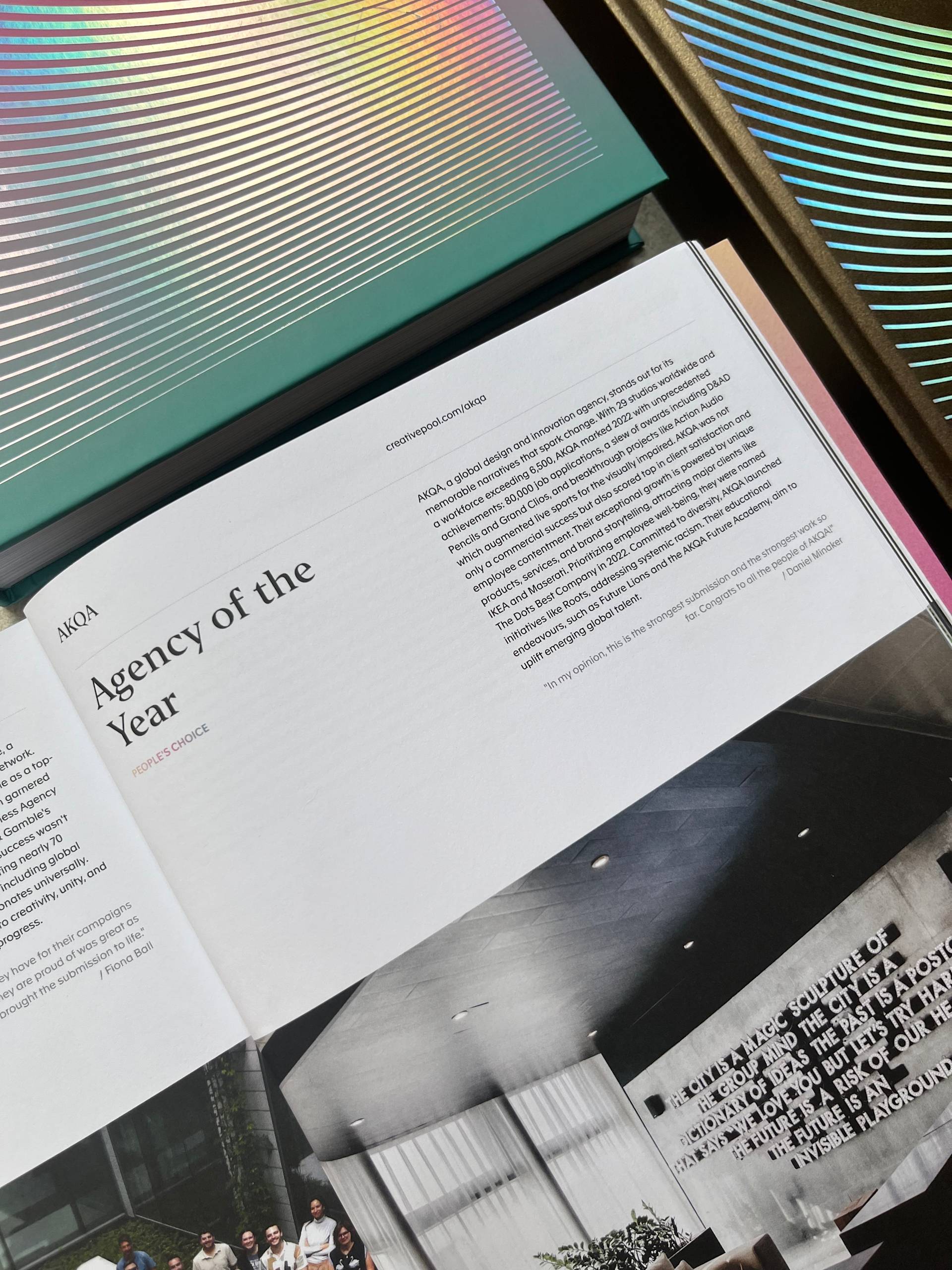 An open magazine spread features an article titled Agency of the Year with a photo of a modern office interior below. Iridescent and colorful book covers are partially visible along the edges of the image.