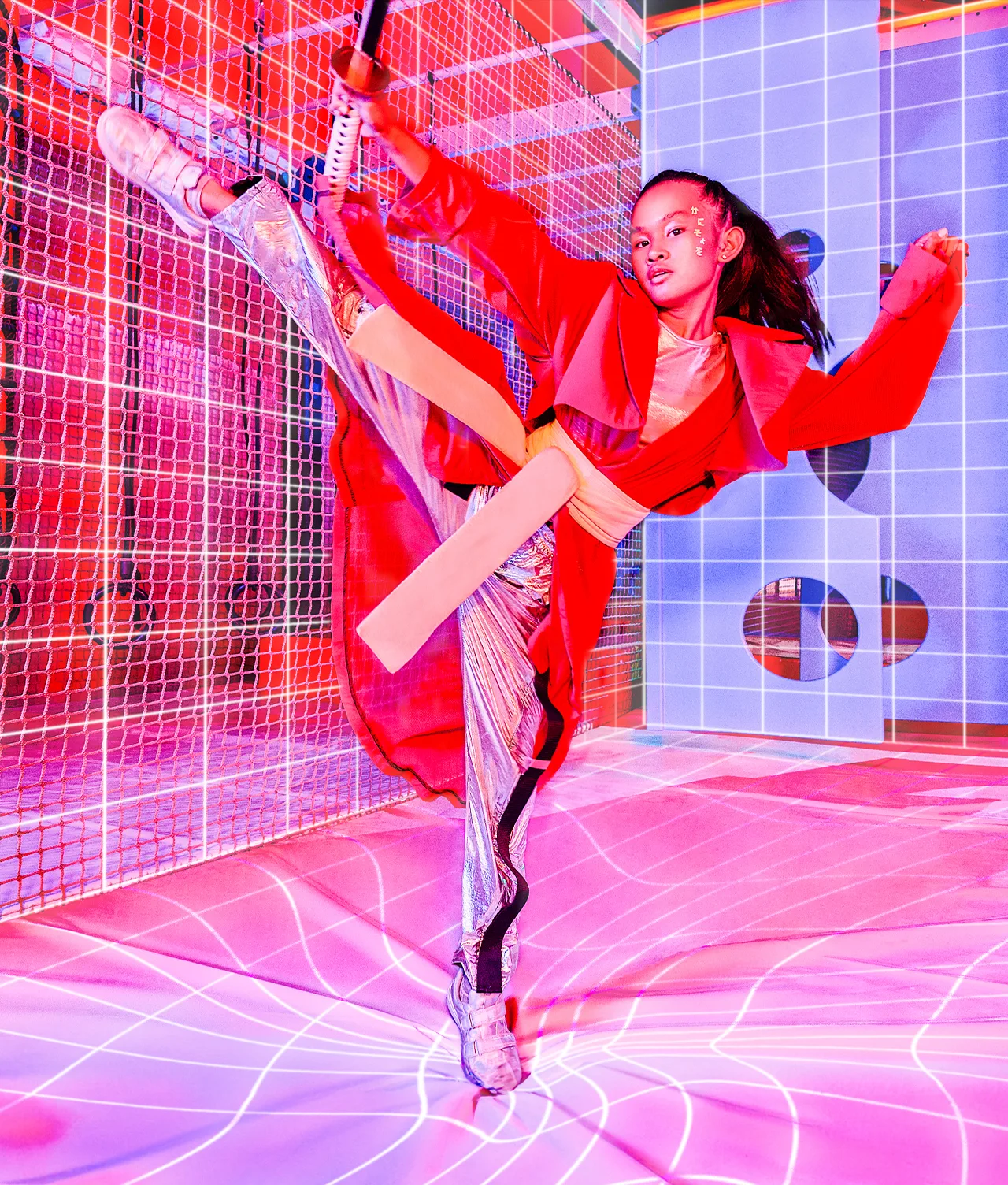 A young woman in futuristic, metallic clothing performs a high kick in a colorful, neon-lit room with grid-patterned walls and floor, holding a staff and surrounded by dramatic pink and blue lighting.