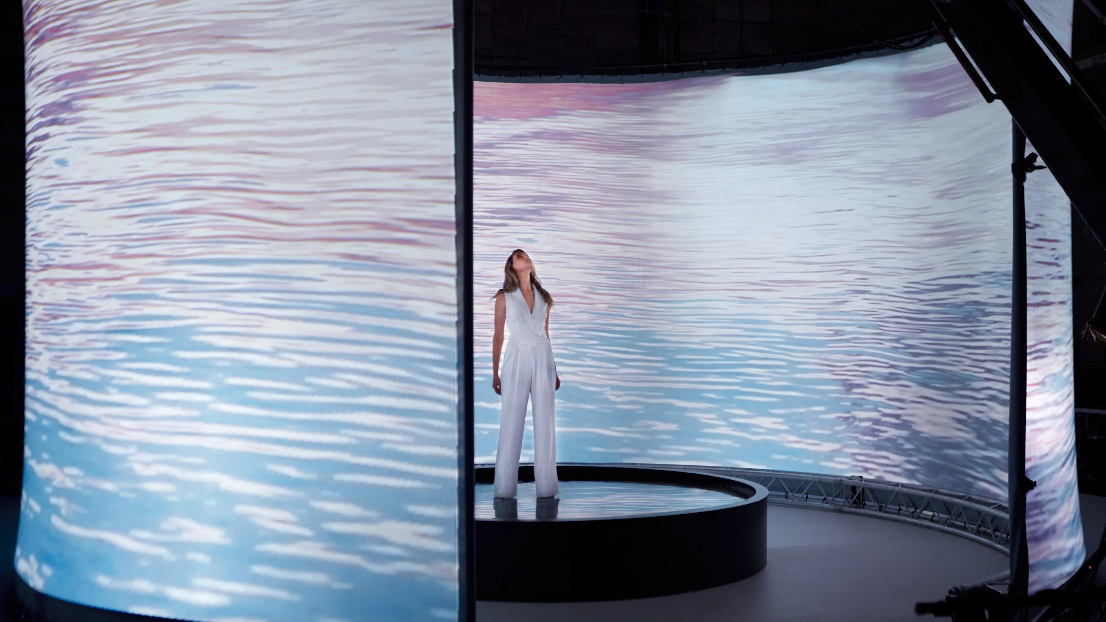 A woman in a white outfit stands on a circular platform surrounded by large curved screens displaying a serene water scene with gentle ripples in pink and blue tones.