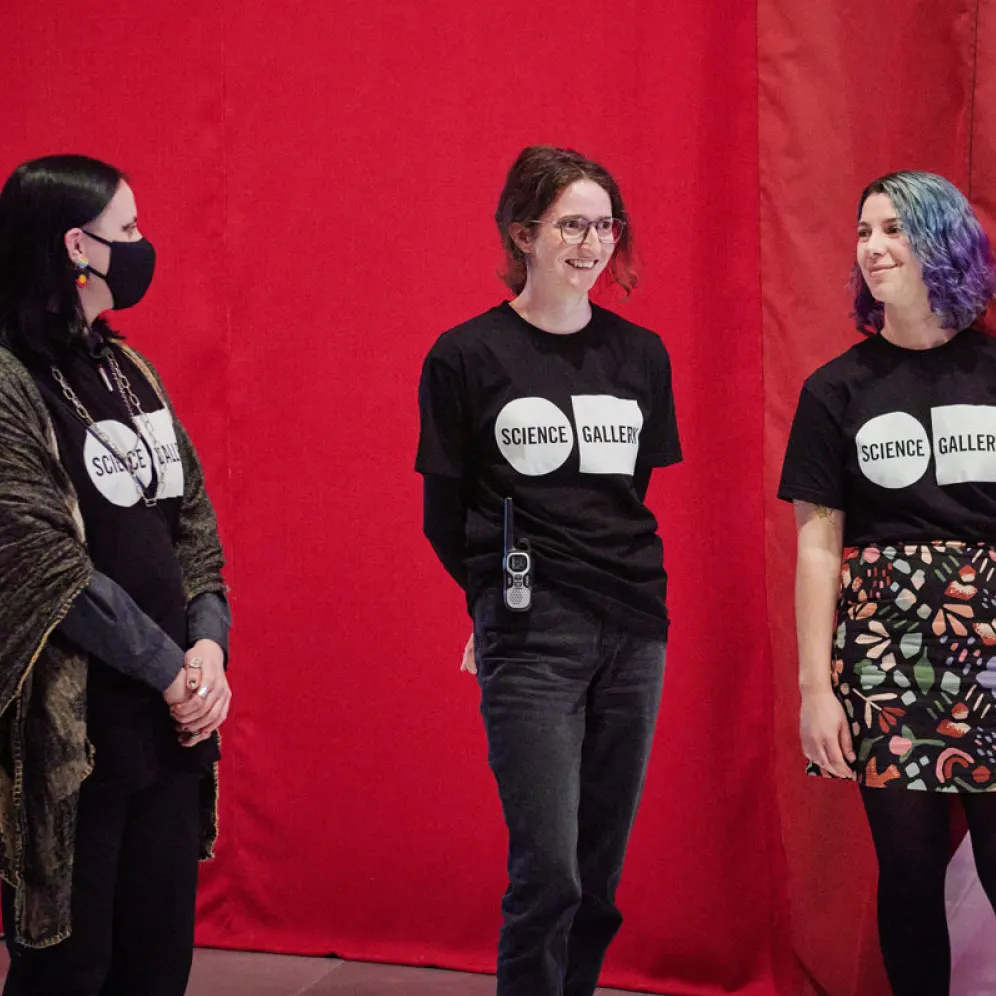 Three people stand in front of a red background, all wearing black Science Gallery t-shirts. One person wears a face mask and a shawl, while the other two are smiling and casually dressed.