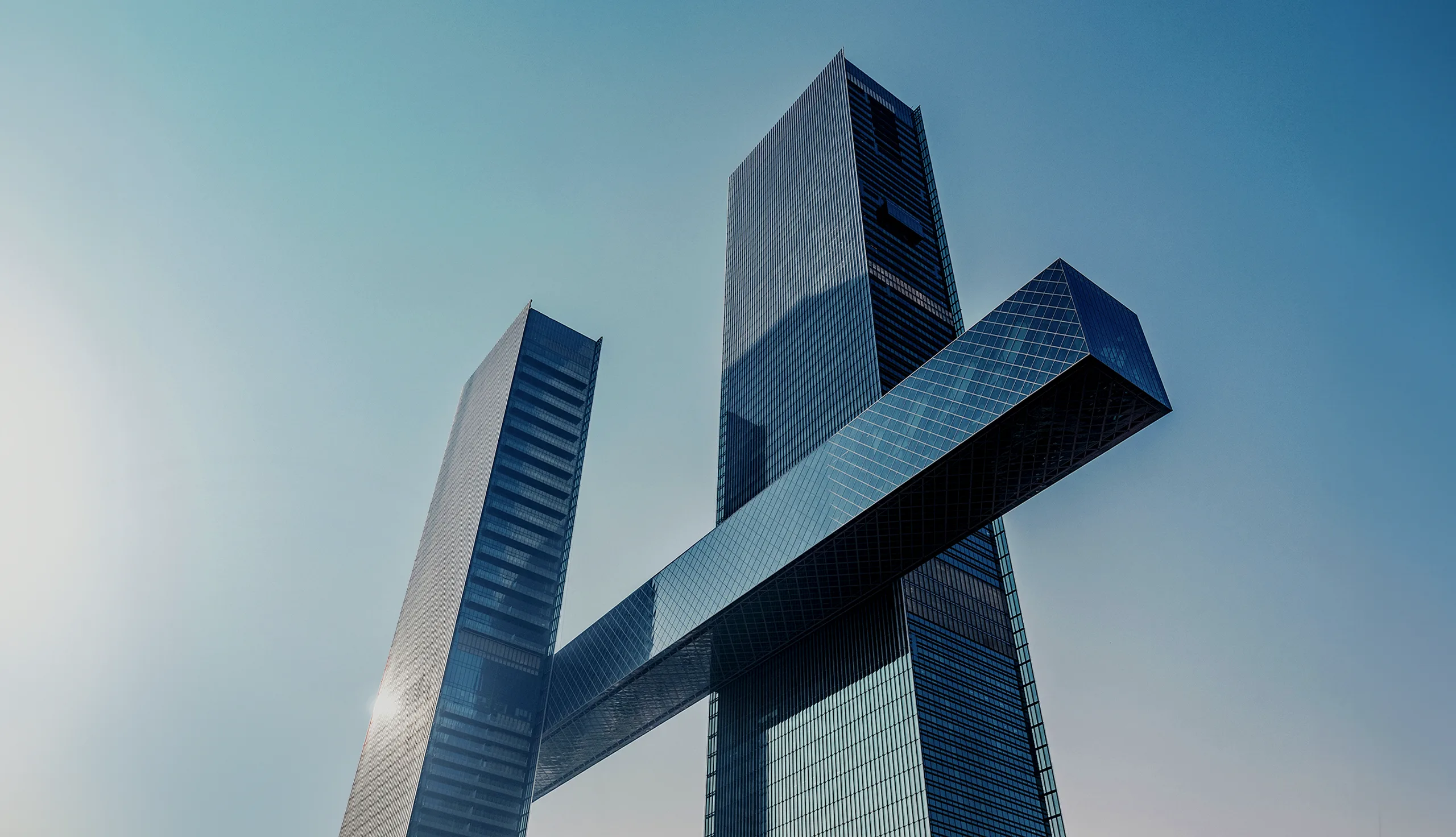 Two modern skyscrapers stand tall against a clear blue sky, with one building featuring a large horizontal structure connecting the towers at a high level. Sunlight reflects off the glass façades.