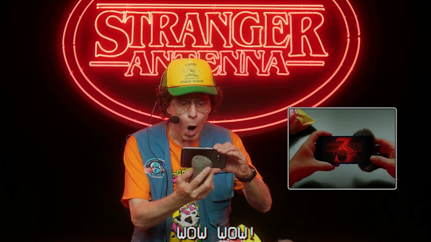 A man in a yellow hat and expressive outfit reacts in shock to a phone, with a Stranger Antenna neon sign behind him. An inset shows hands holding a phone displaying 3 and TV static. Subtitles read WOW WOW!.