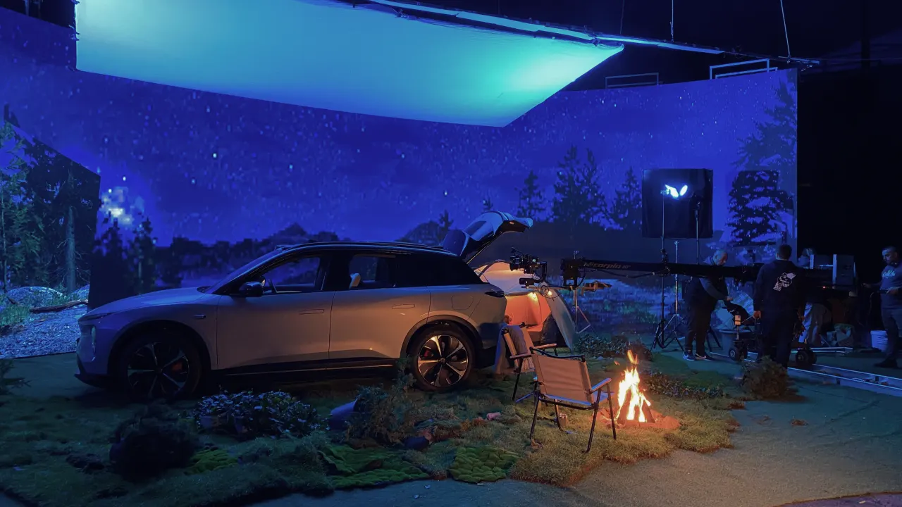 A car is parked on a set designed to look like a nighttime campsite, with artificial grass, a campfire, chairs, and a scenic forest backdrop. Several people and studio lights are visible in the background.