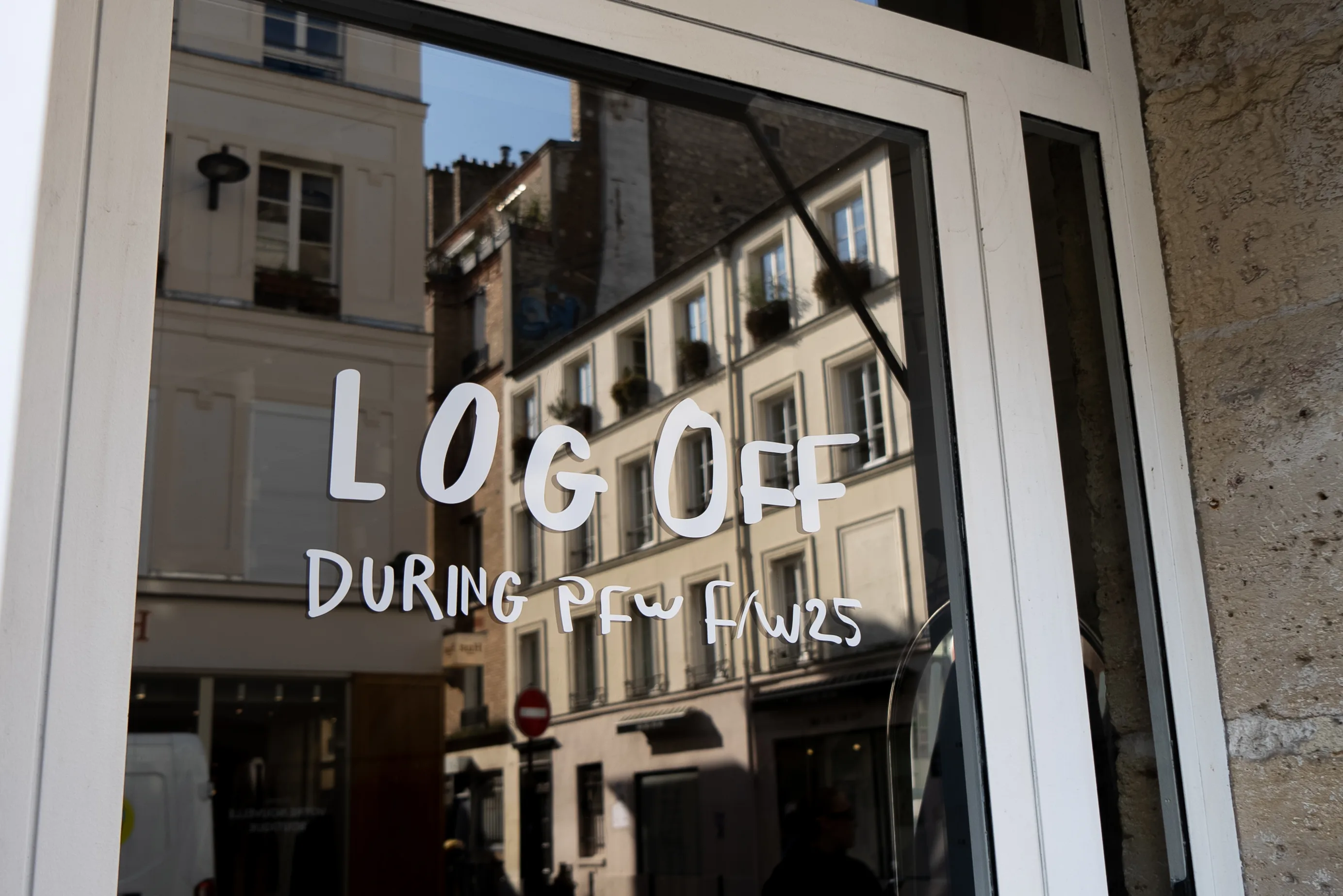 A glass door with the handwritten message LOG OFF DURING PFW FW25 in white letters, reflecting nearby Parisian buildings and a street scene.