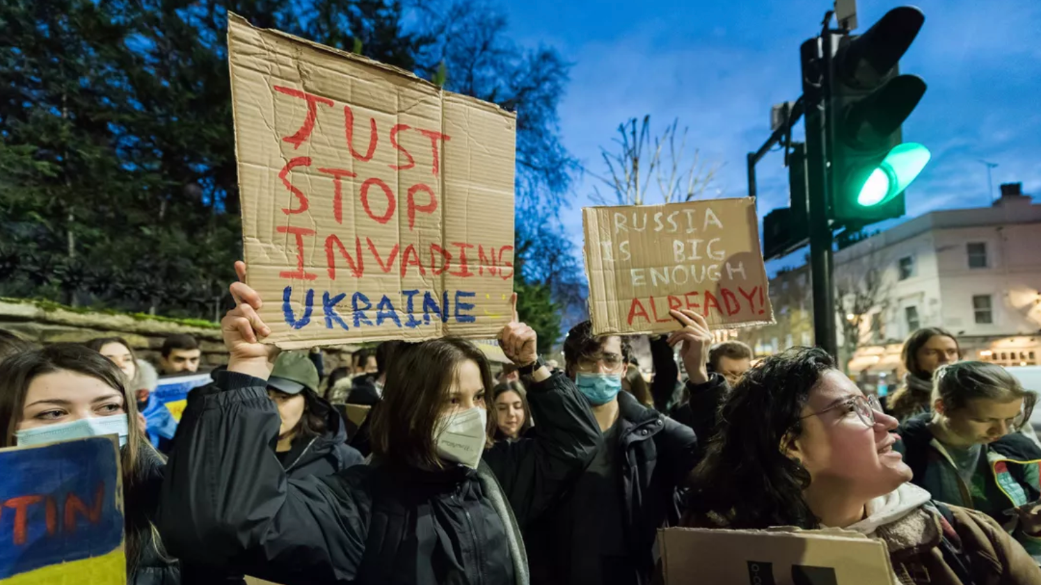 A group of protesters, some wearing masks, hold cardboard signs reading JUST STOP INVADING UKRAINE and RUSSIA IS BIG ENOUGH ALREADY at a street demonstration in the evening.